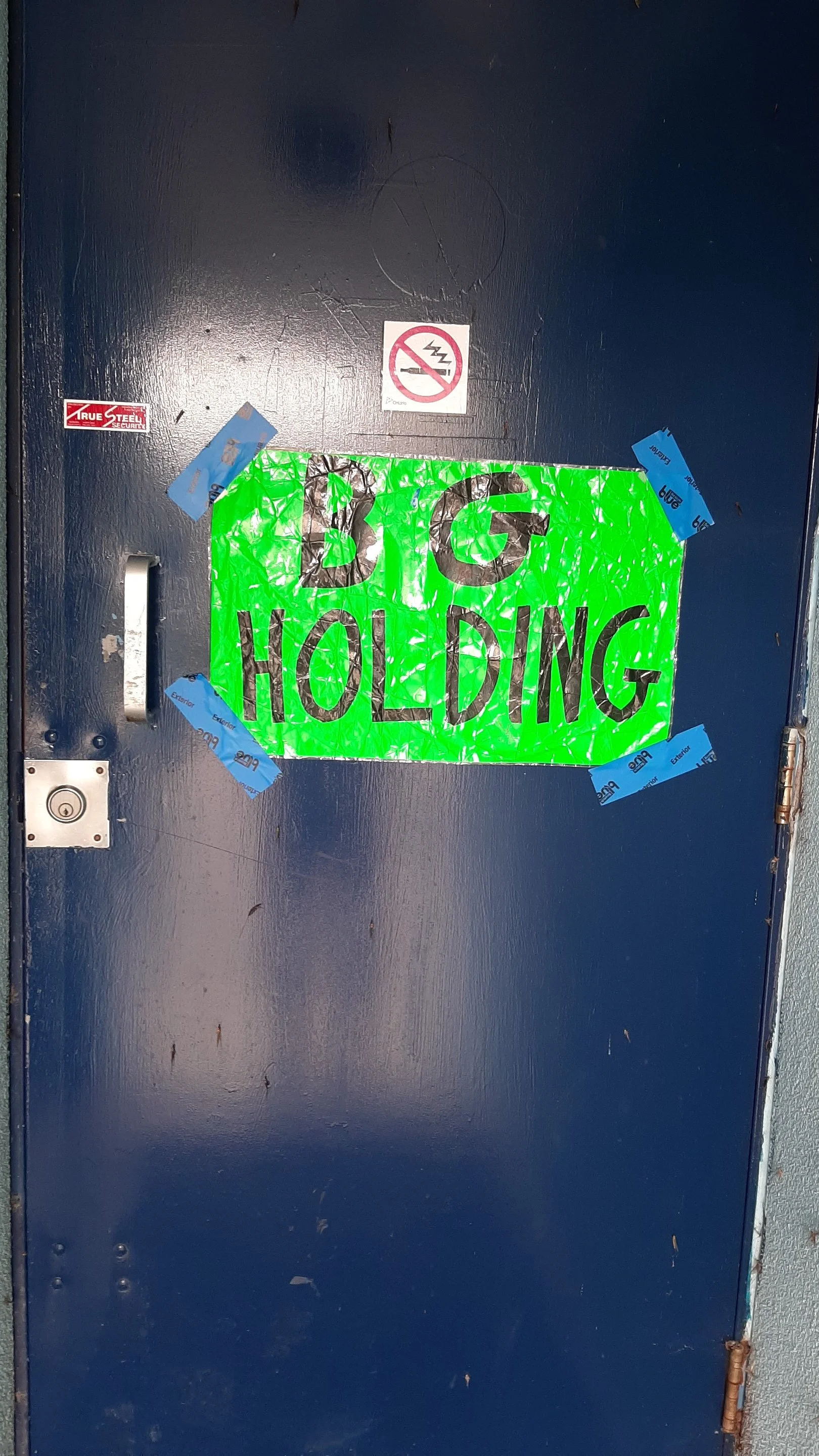 Bright green sign on a blue door that says 'BIG HOLDING' in black letters, taped with blue painter's tape along the edges.
