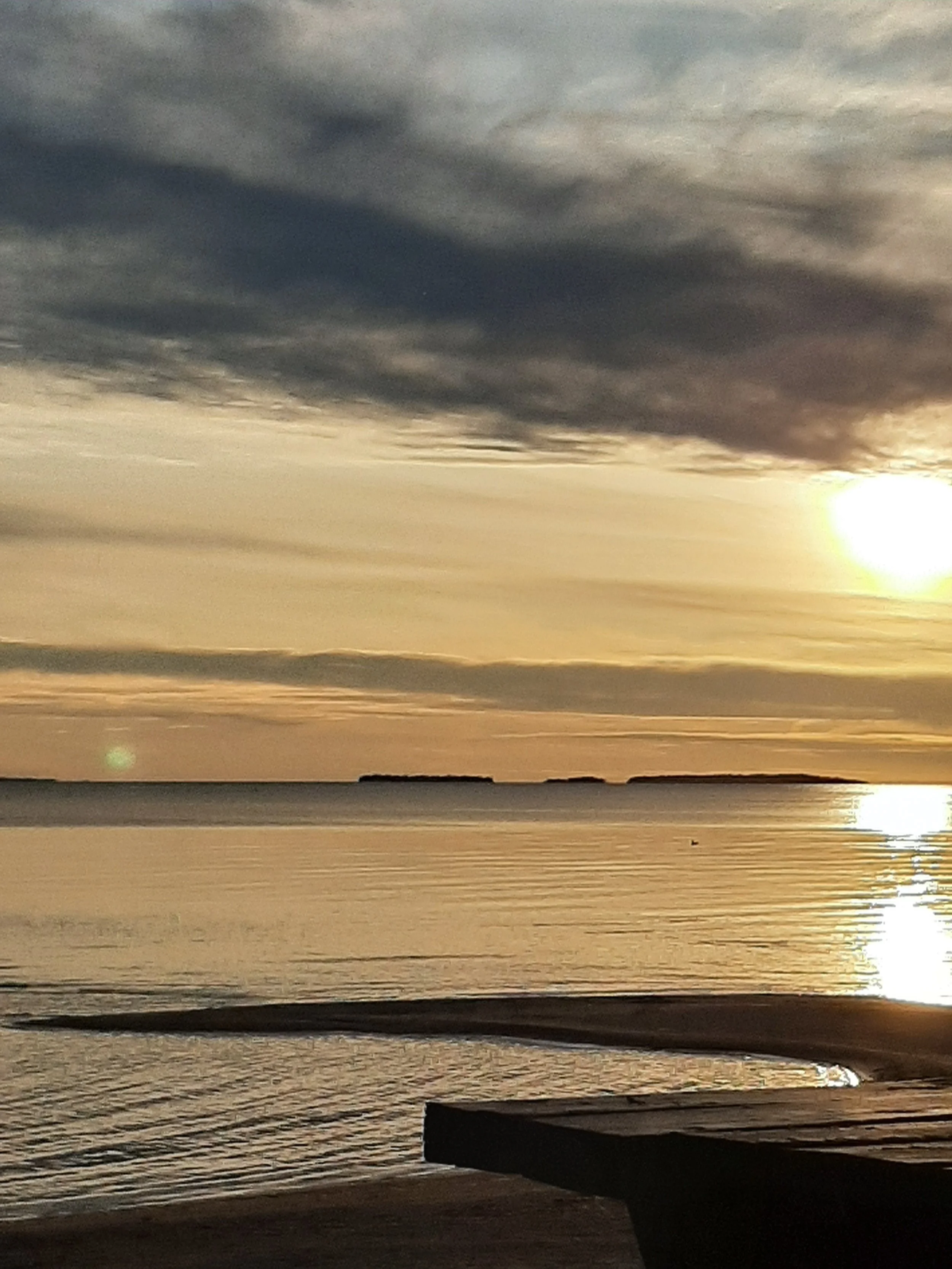 Sunset over a calm ocean with dark clouds overhead and a silhouette of an island in the distance.