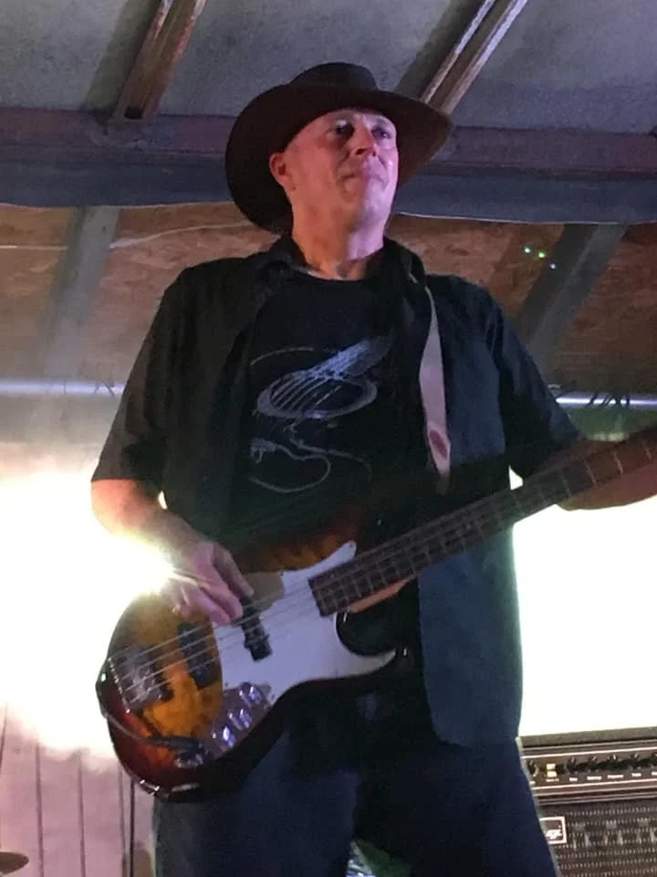 A man wearing a black cowboy hat and a black shirt playing an electric bass guitar on stage with purple lighting.