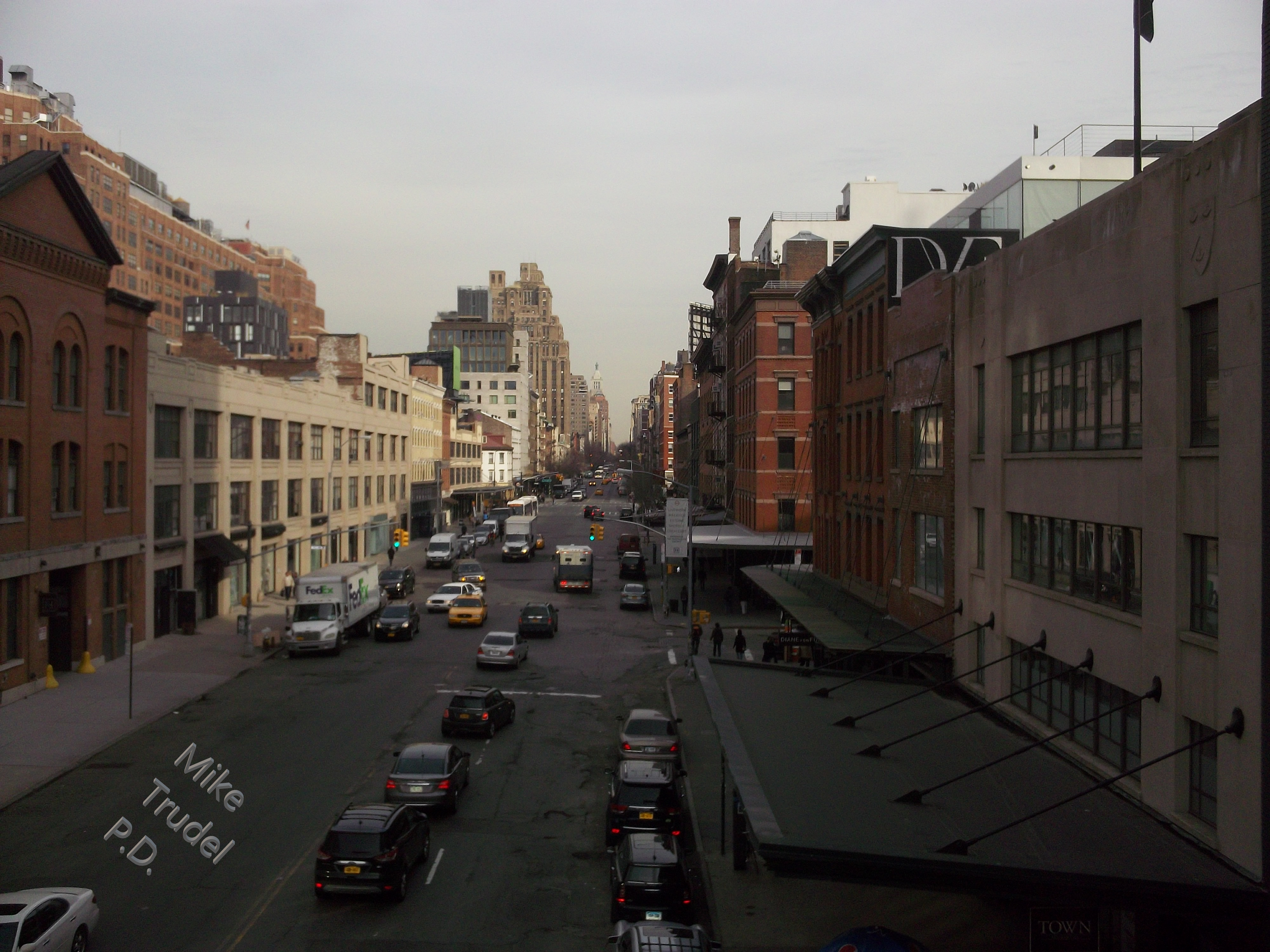 City street view with cars and buildings on both sides, overcast sky, with the text 'Mike Trudel P.D.' on the street and a large building with a sign "D" visible on the right.