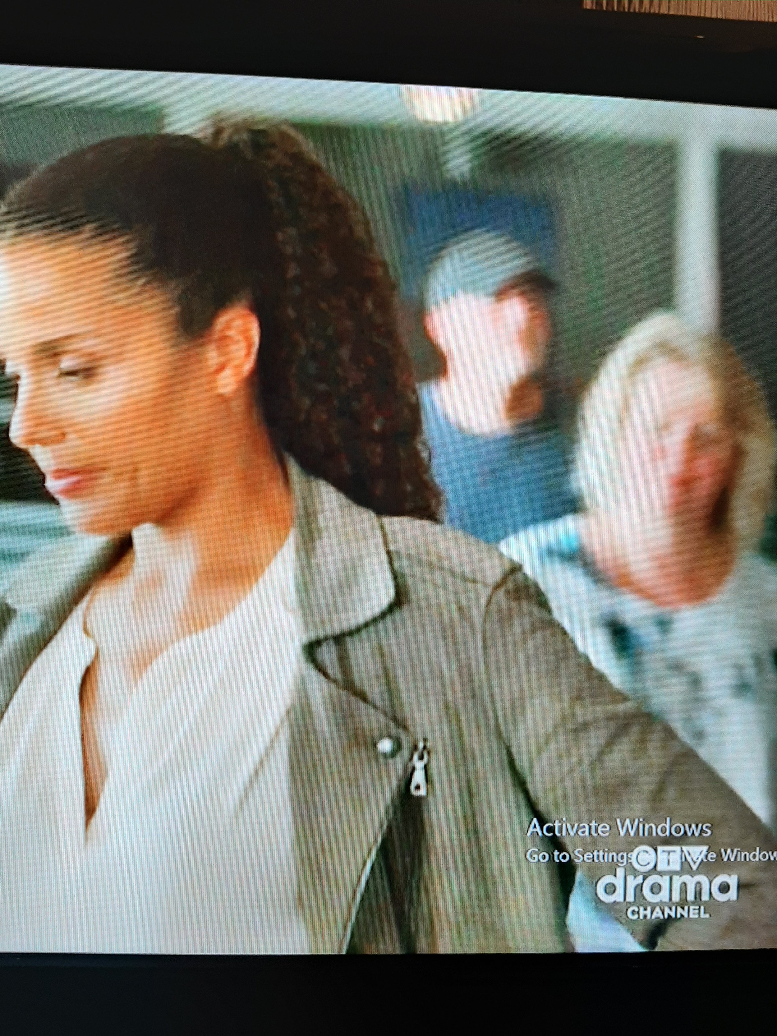 Close-up of a woman with dark curly hair in a ponytail, wearing a tan jacket, with two blurred people in the background, one man with a hat and a woman with blonde hair, inside a building.