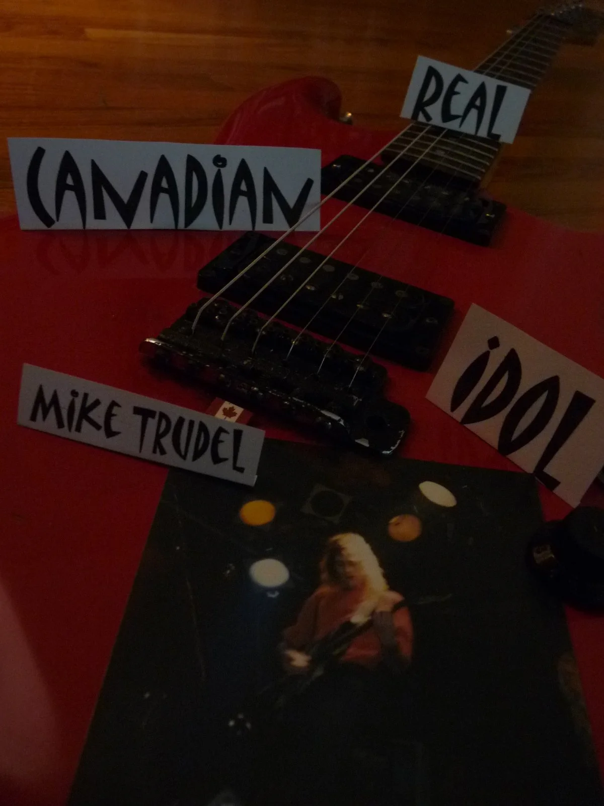 A red electric guitar with handwritten signs that say "Canadian," "Real," "Idol," and "Mike Trudel." There is also a photo of a woman playing a bass guitar, with colored lights in the background.