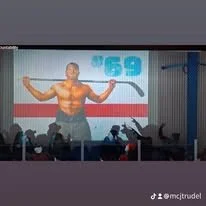 A shirtless man standing on stage with arms outstretched, speaking into a microphone with the number '69' displayed on a screen behind him.