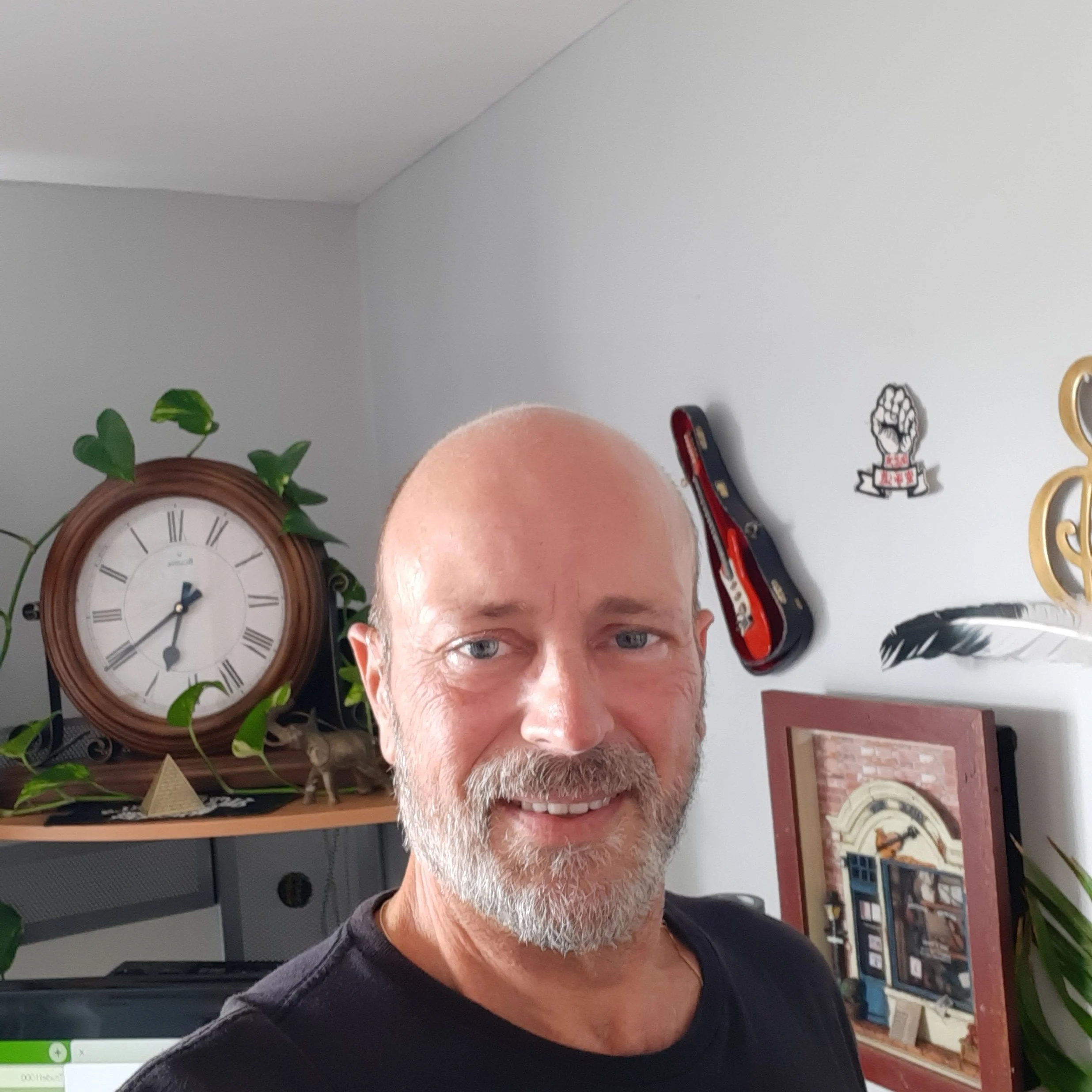 Smiling man with a white beard and crew cut, standing in a room with a clock, guitars, and artwork on the wall.