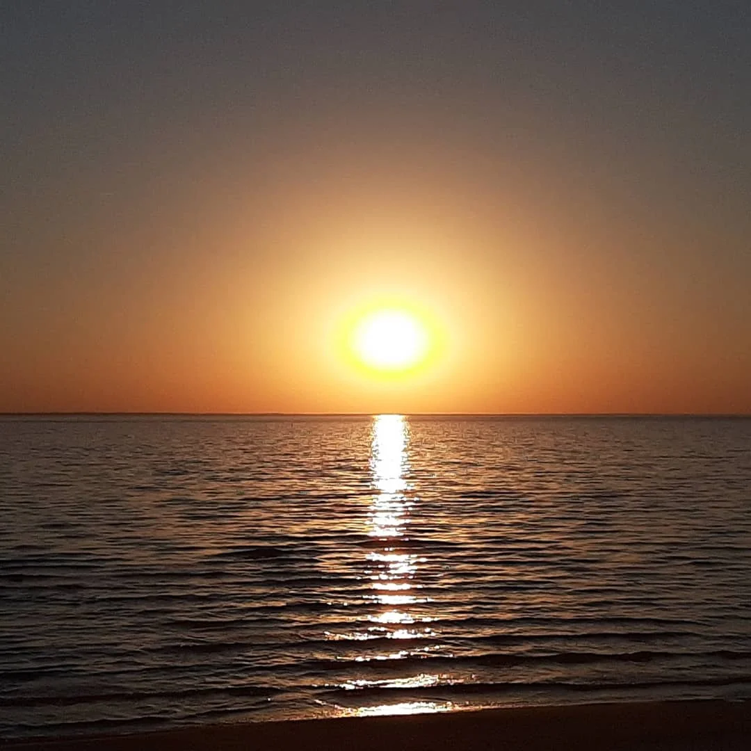 Sunset over the ocean with a reflection on the water and a sandy beach in the foreground.