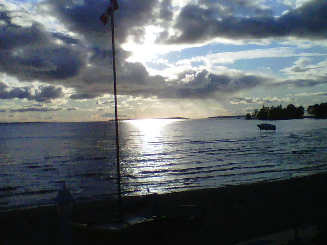 A lake scene with a cloudy sky and the sun reflecting on the water, with a small boat on the shore and trees in the distance.