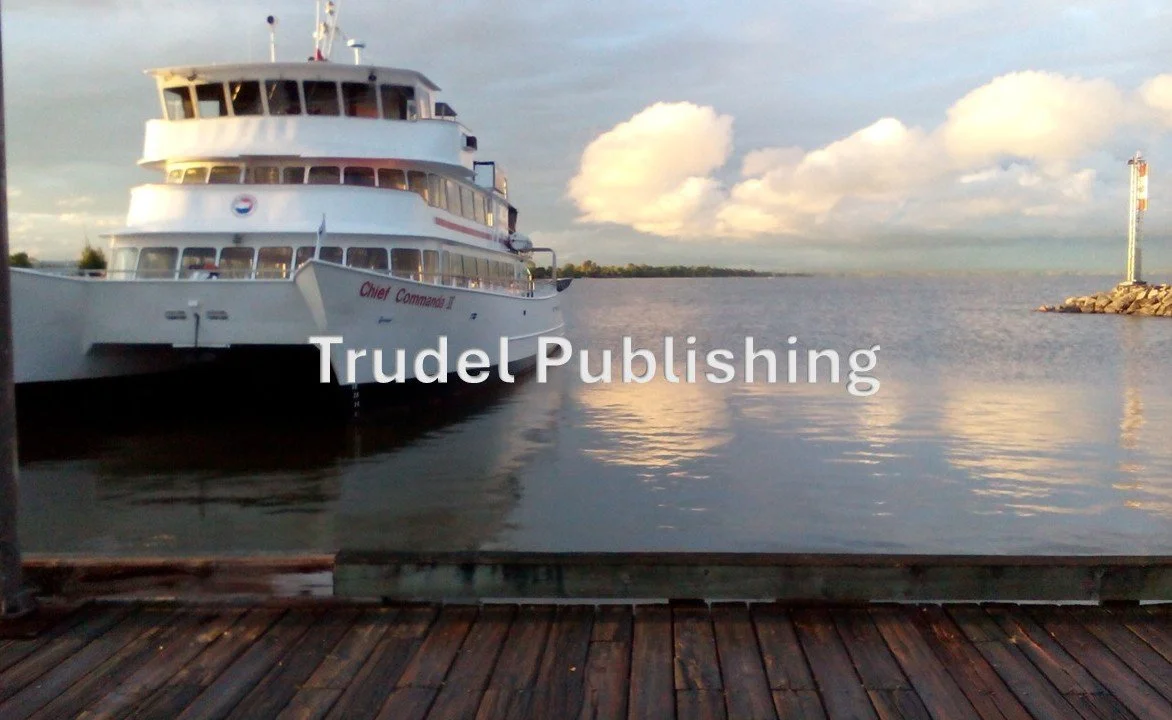 A large white boat docked at a wooden pier on a body of water, with clouds in the sky and a lighthouse in the distance.