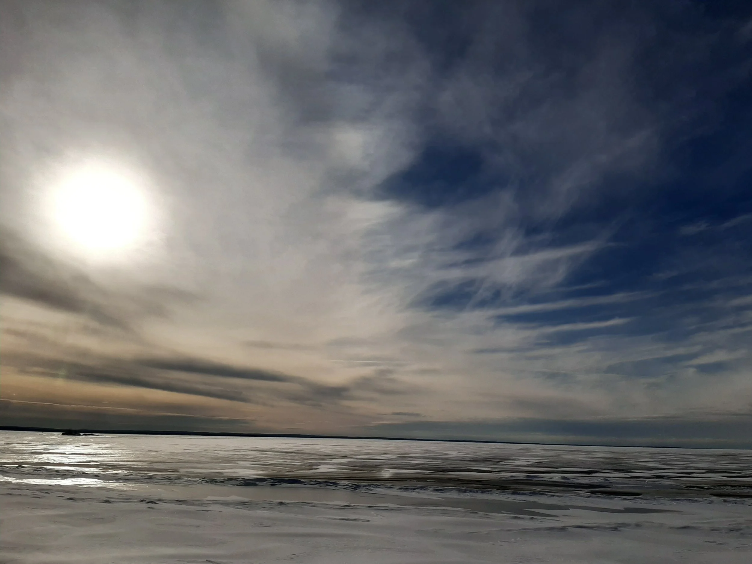 A winter landscape with a snow-covered ground, a frozen lake, and an overcast sky with the sun partially visible through the clouds.