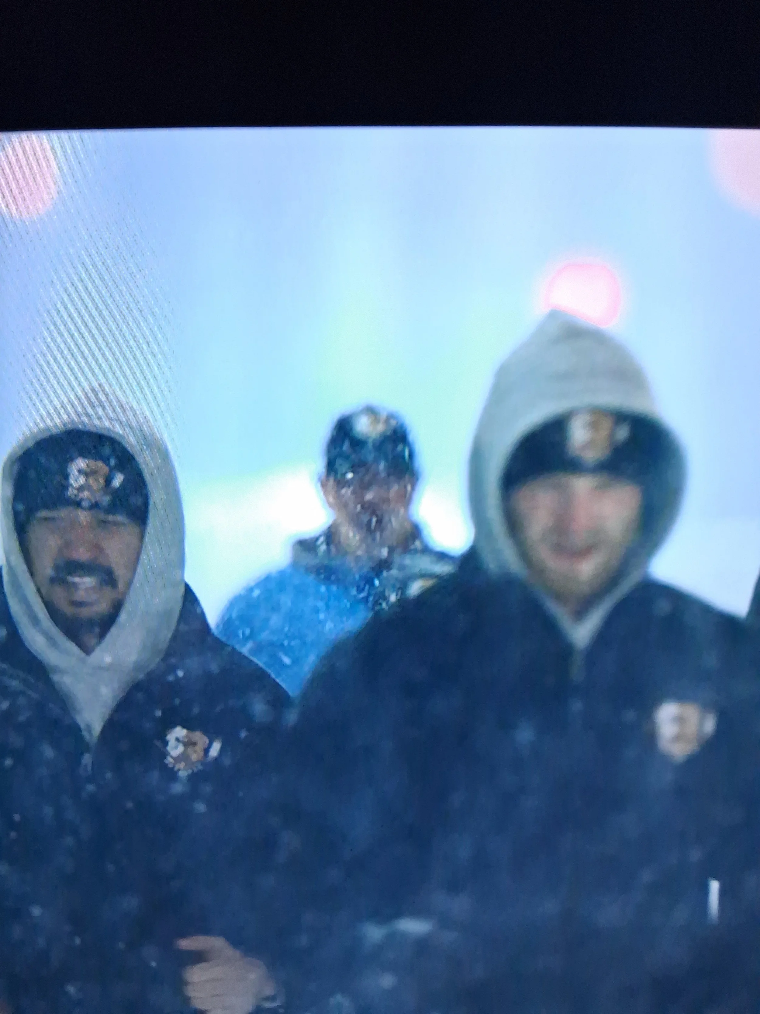 Three men in black hoodies with gray hoods, two with airplane patches on their caps, standing in front of a light blue background, with raindrops or water splashes visible.