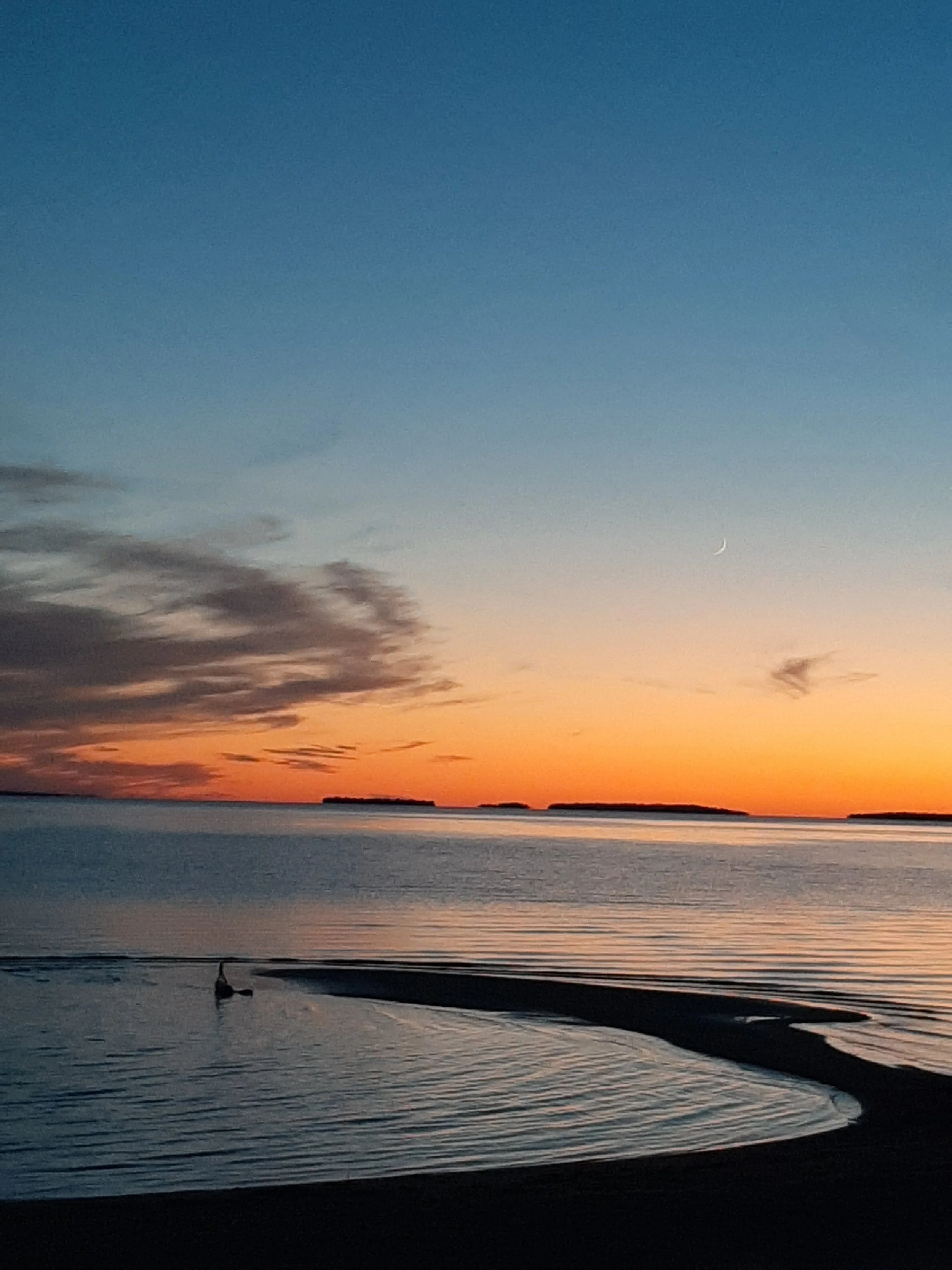 Sunset over a calm body of water with a small sandy shore in the foreground, a person swimming, and islands on the horizon, with a crescent moon in the sky.