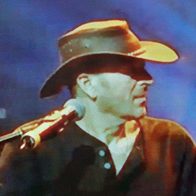 A man wearing a cowboy hat and black shirt, positioned near a microphone, against a dark background.