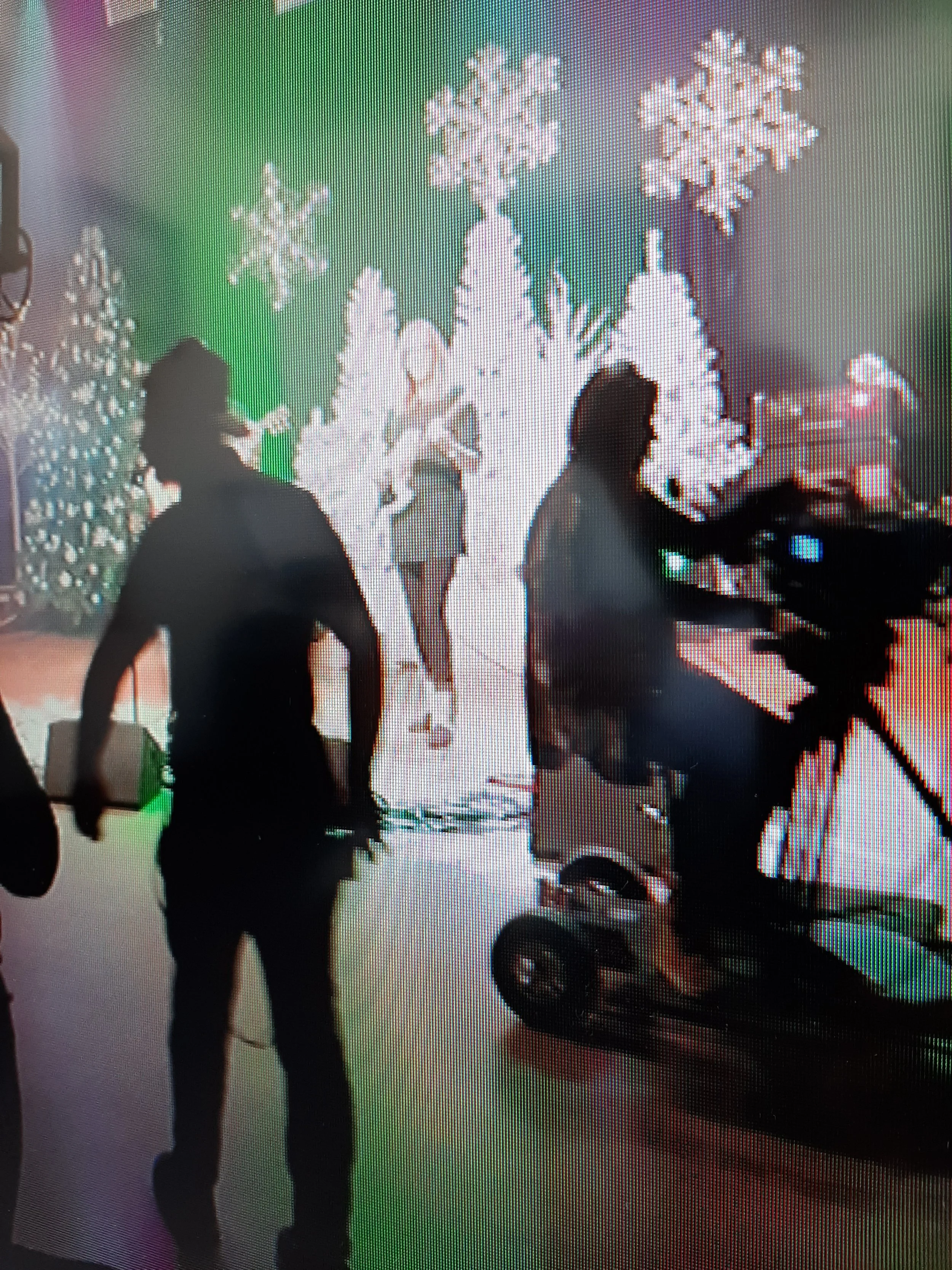 Behind the scenes of a holiday photo shoot with white Christmas trees decorated with snowflake ornaments, and a girl in a gray dress in front of the trees, with crew members setting up equipment.