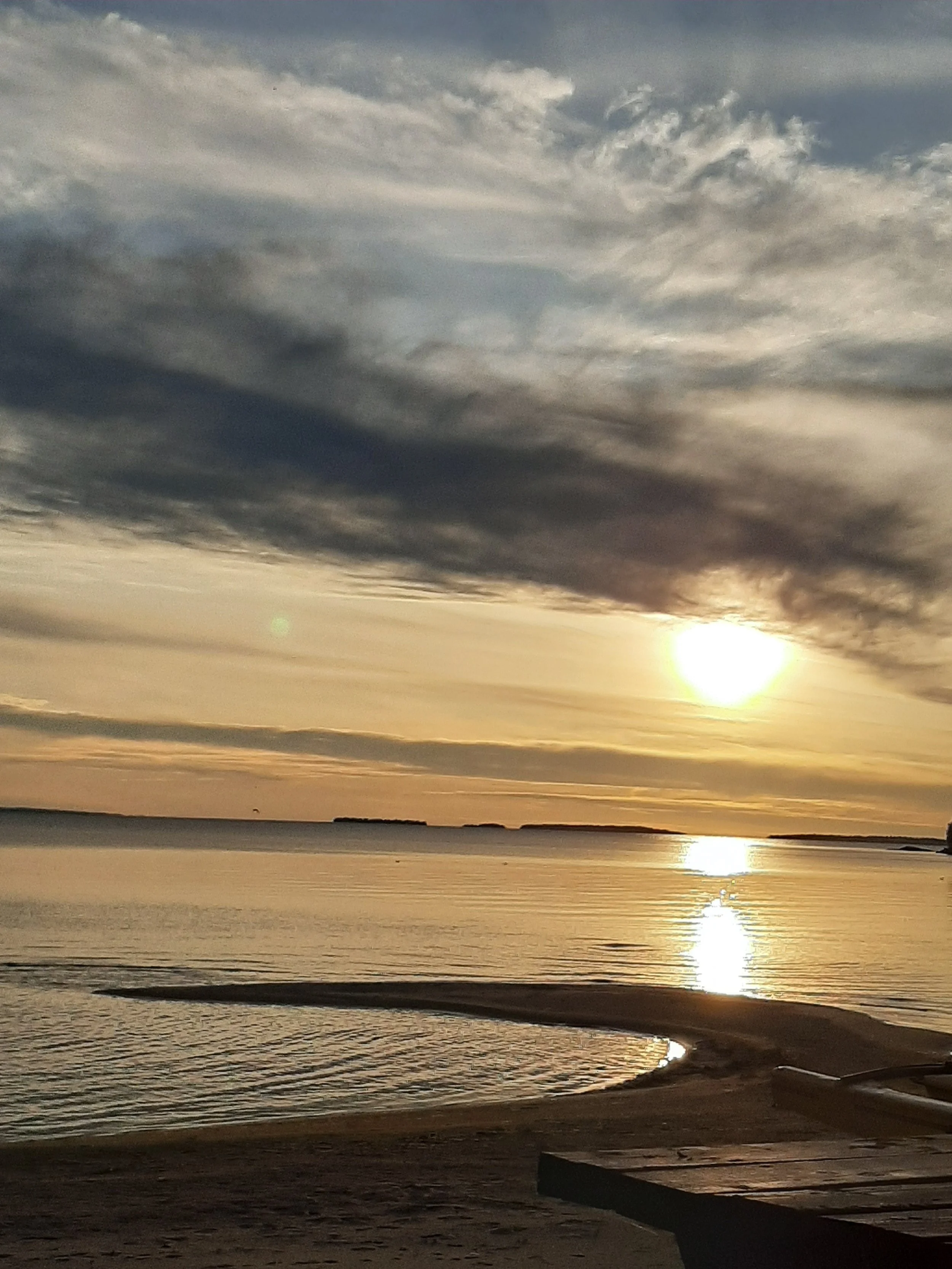 A beach at sunset with a cloudy sky, calm water reflecting the sunlight, and a sandy shoreline with a small concrete structure.