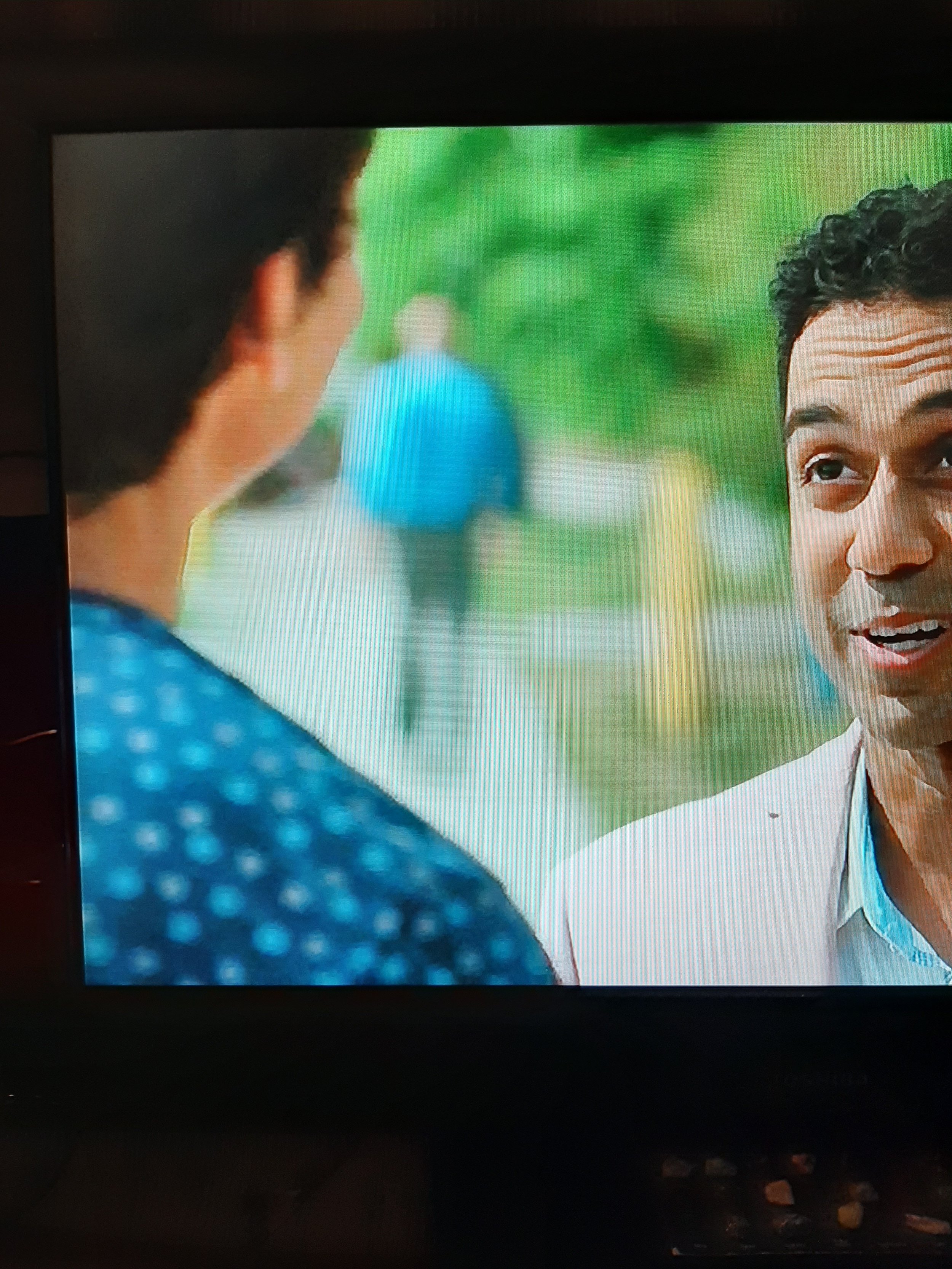Close-up of two men talking outside, one facing the camera smiling, the other with his back to the camera in a blue shirt, with blurred greenery and a person walking in the background.