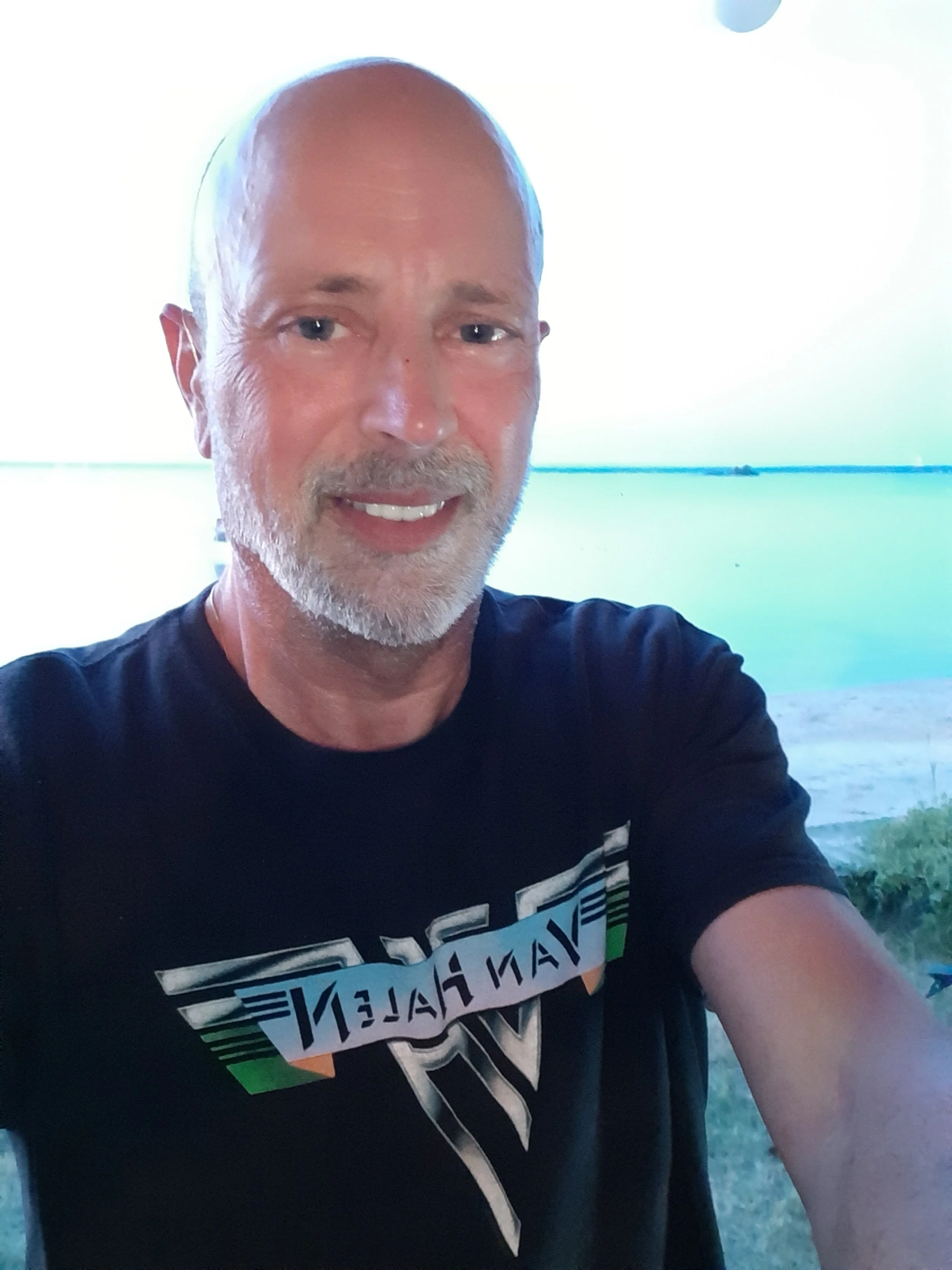 A middle-aged man with a beard and shaved head taking a selfie outdoors at a beach or lakeside during daytime, with water in the background.