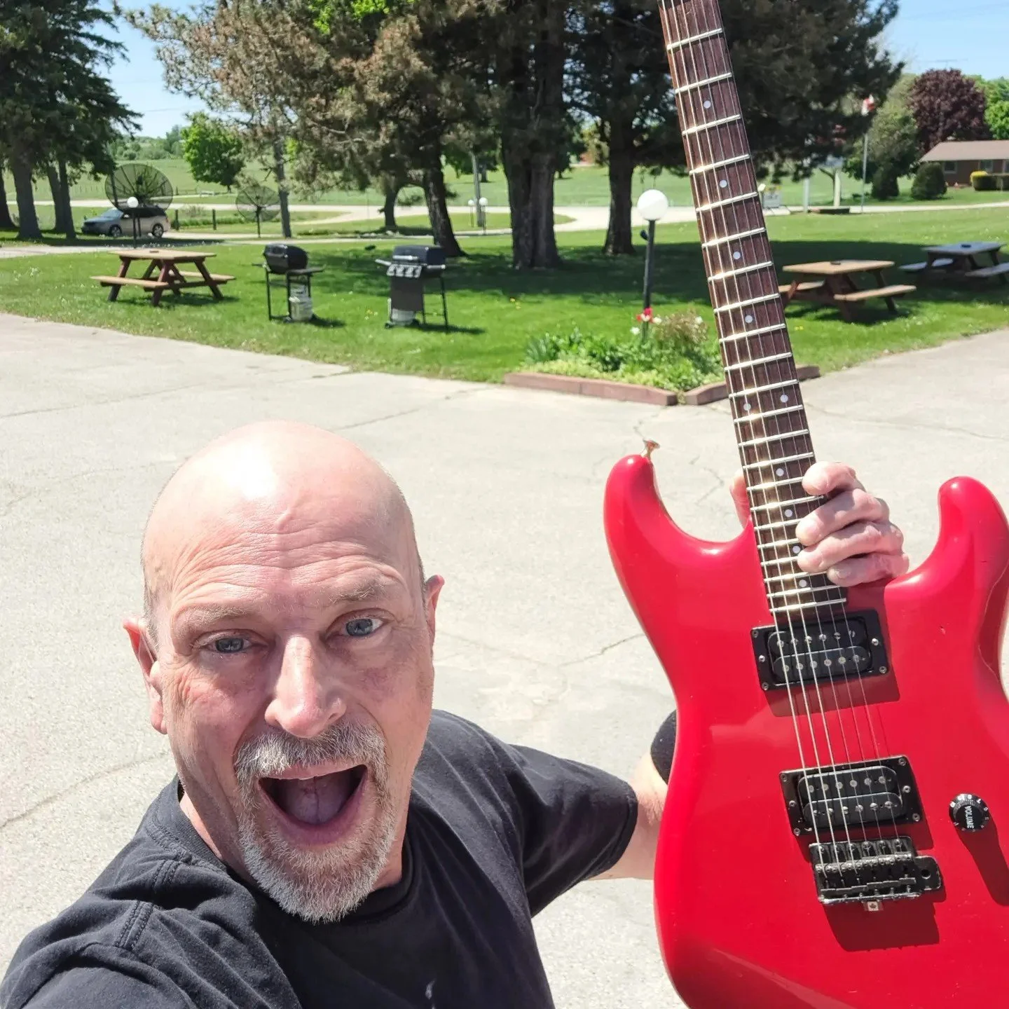 A man with a bald head and gray beard, smiling excitedly, holding a red electric guitar outdoors on a sunny day.
