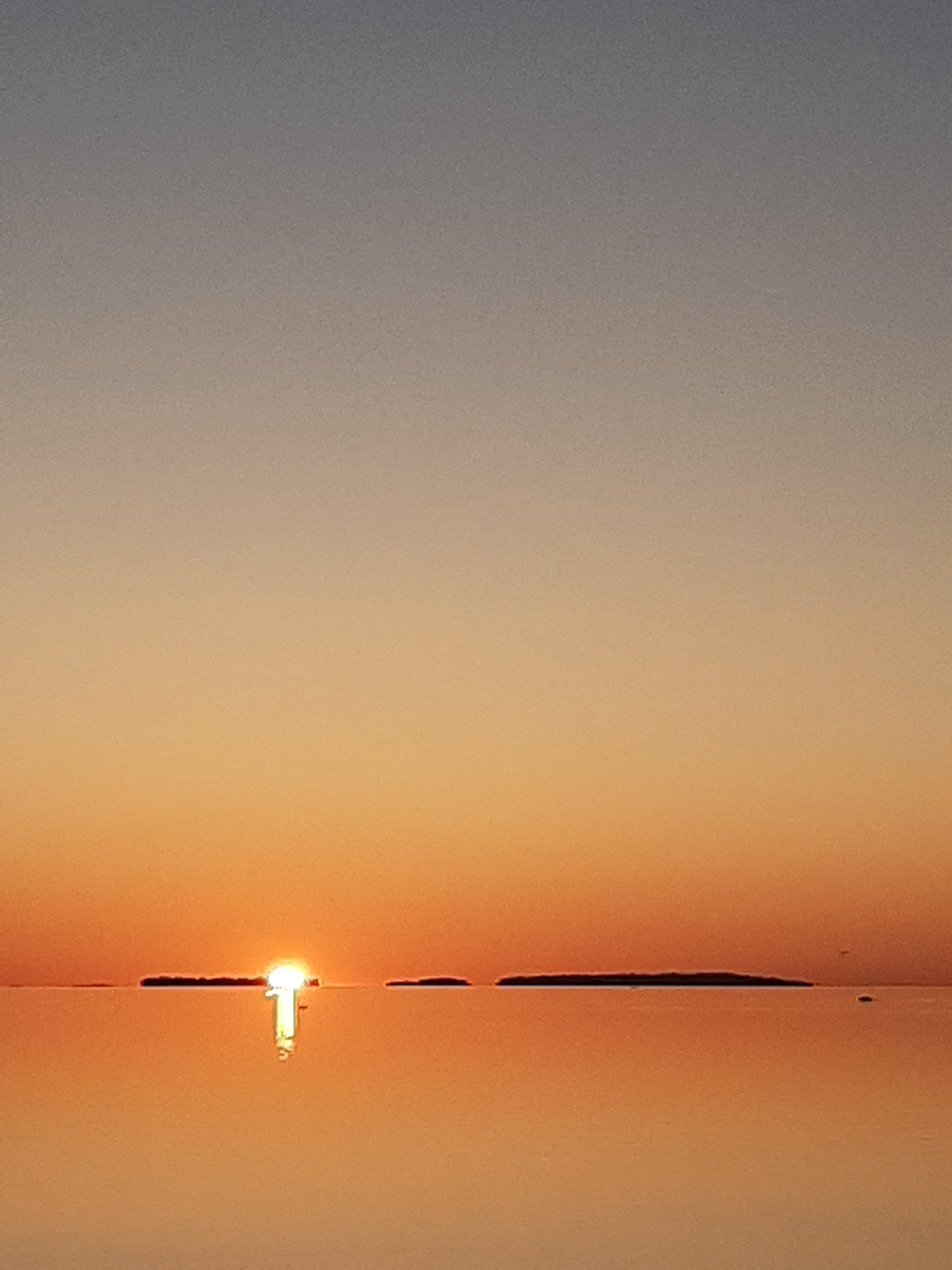 Sun setting over water with small islands in the distance, casting a reflection on the water's surface.