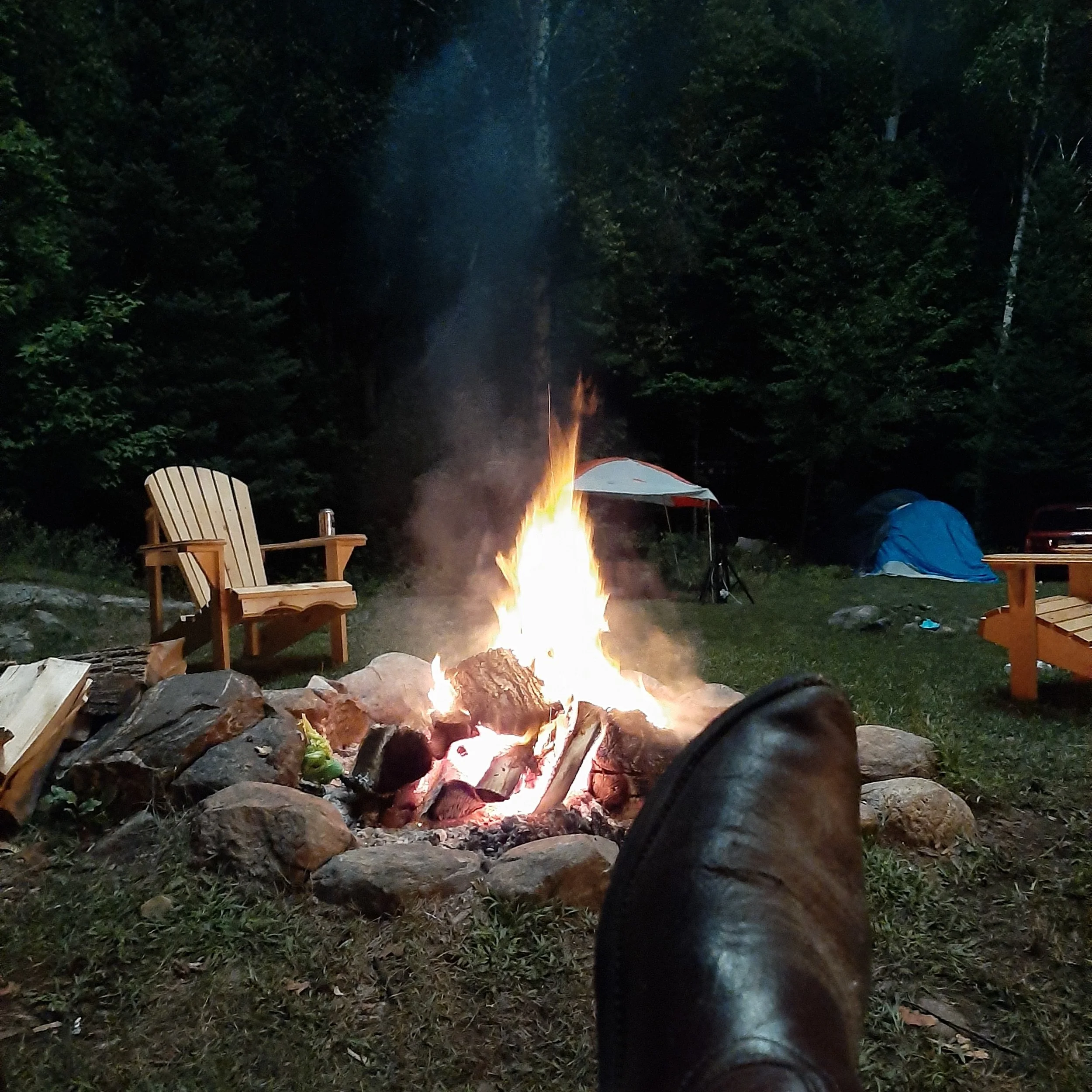 A campfire surrounded by rocks with adirondack chairs and camping tents in a forest at night.