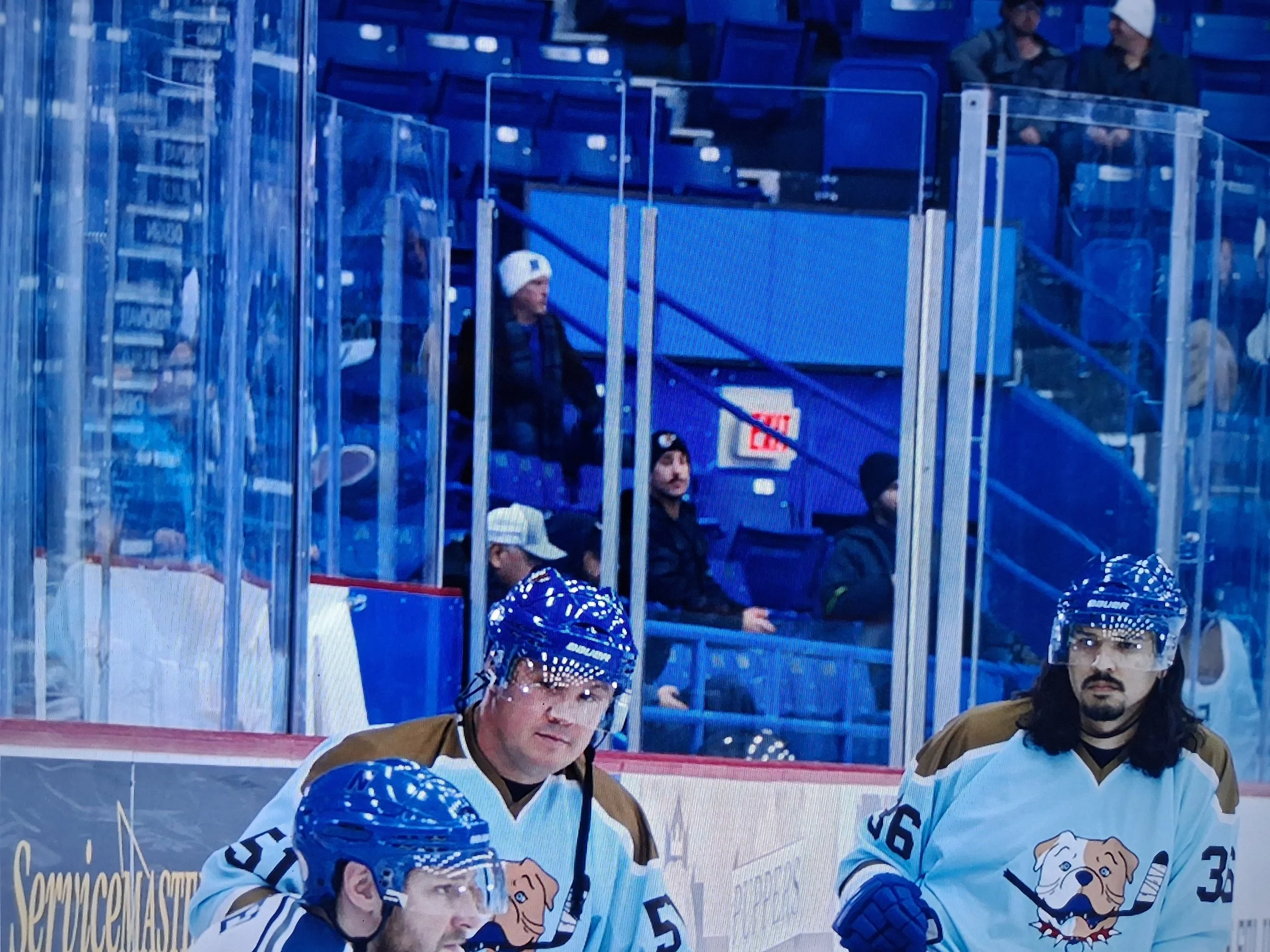Ice hockey players on the ice rink, wearing white jerseys with a bulldog logo, and blue helmets, with some spectators sitting behind the glass barriers in the stands.