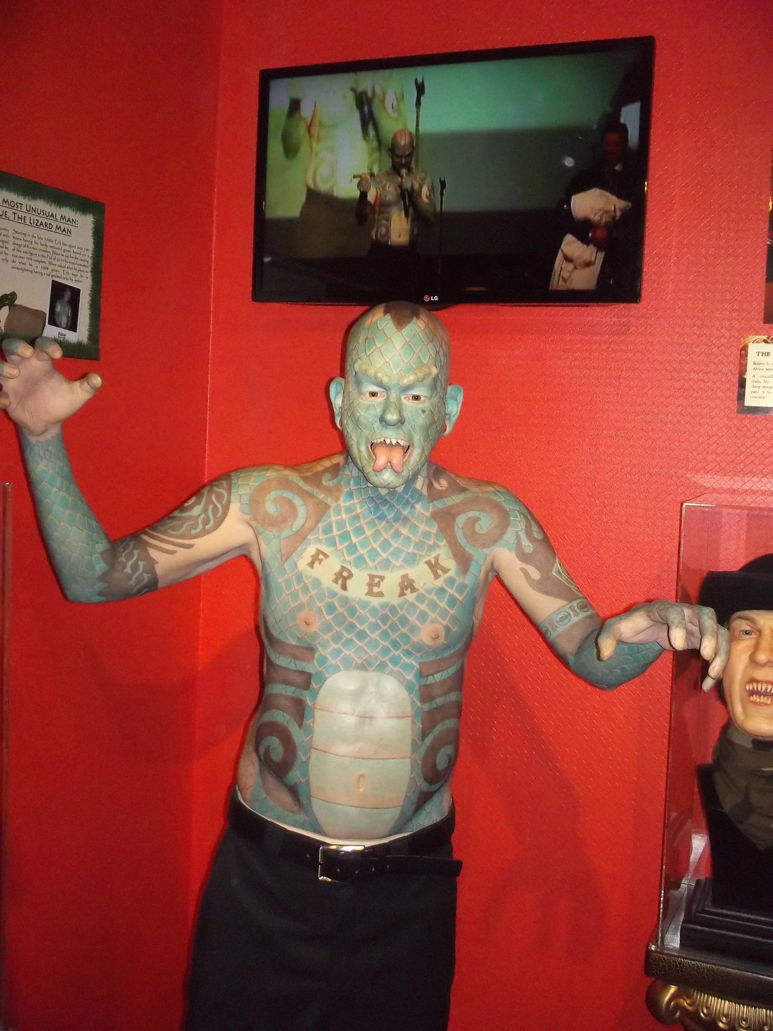 A person with extensive tattoos on upper body, face, and arms poses with arms raised. The tattoos include a dragon or snake pattern, the word 'FREAK' across the chest, and a muscular torso. The individual has a painted face resembling a monster or creature with an open mouth, sharp teeth, and a reptilian or scaly appearance. The background is red, and a screen above shows a man in a similar costume speaking into a microphone.