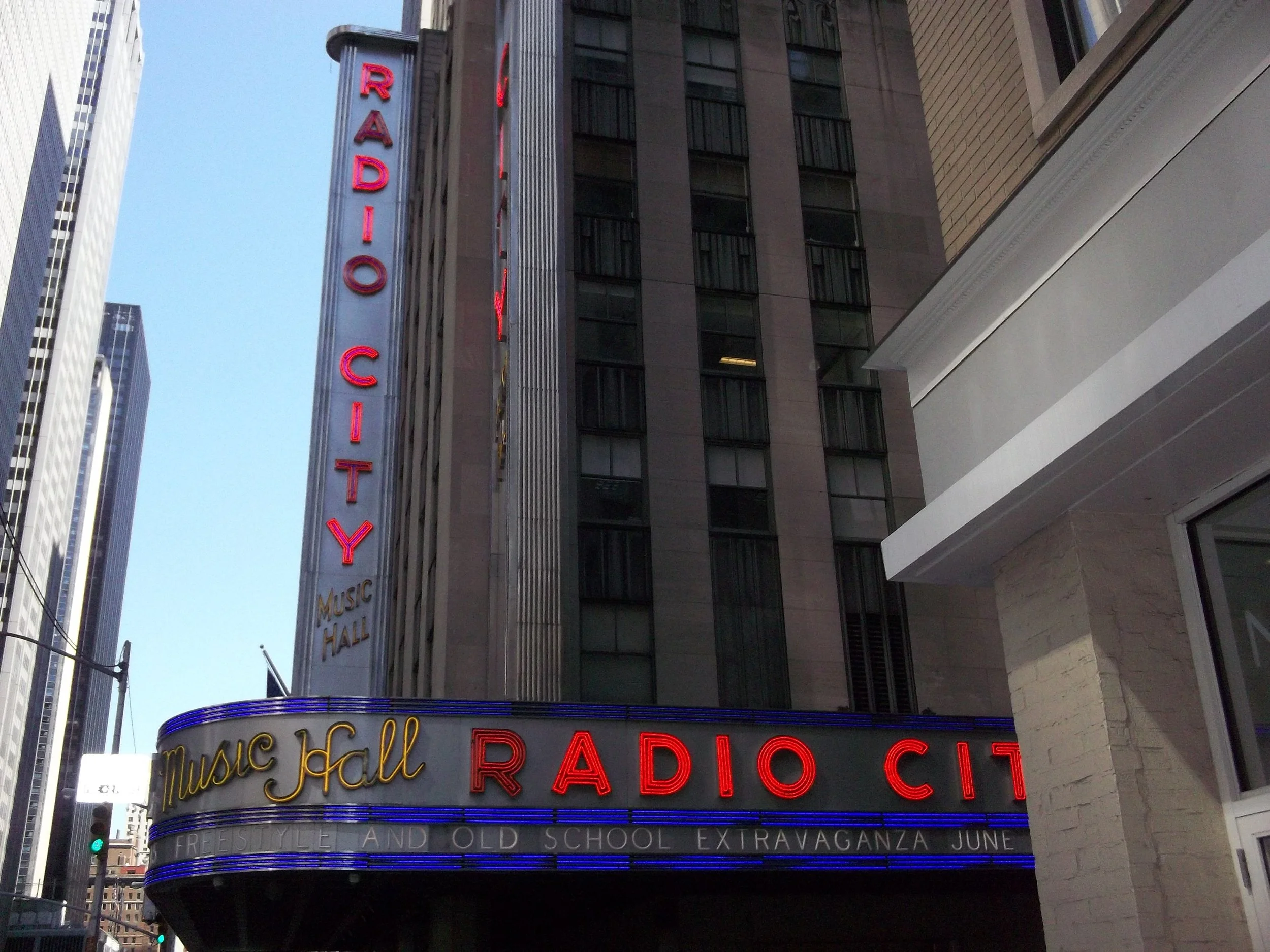 The marquee of Radio City Music Hall with neon lights and vertical sign, displaying the venue's name in red and gold colors, located in an urban area with tall buildings.