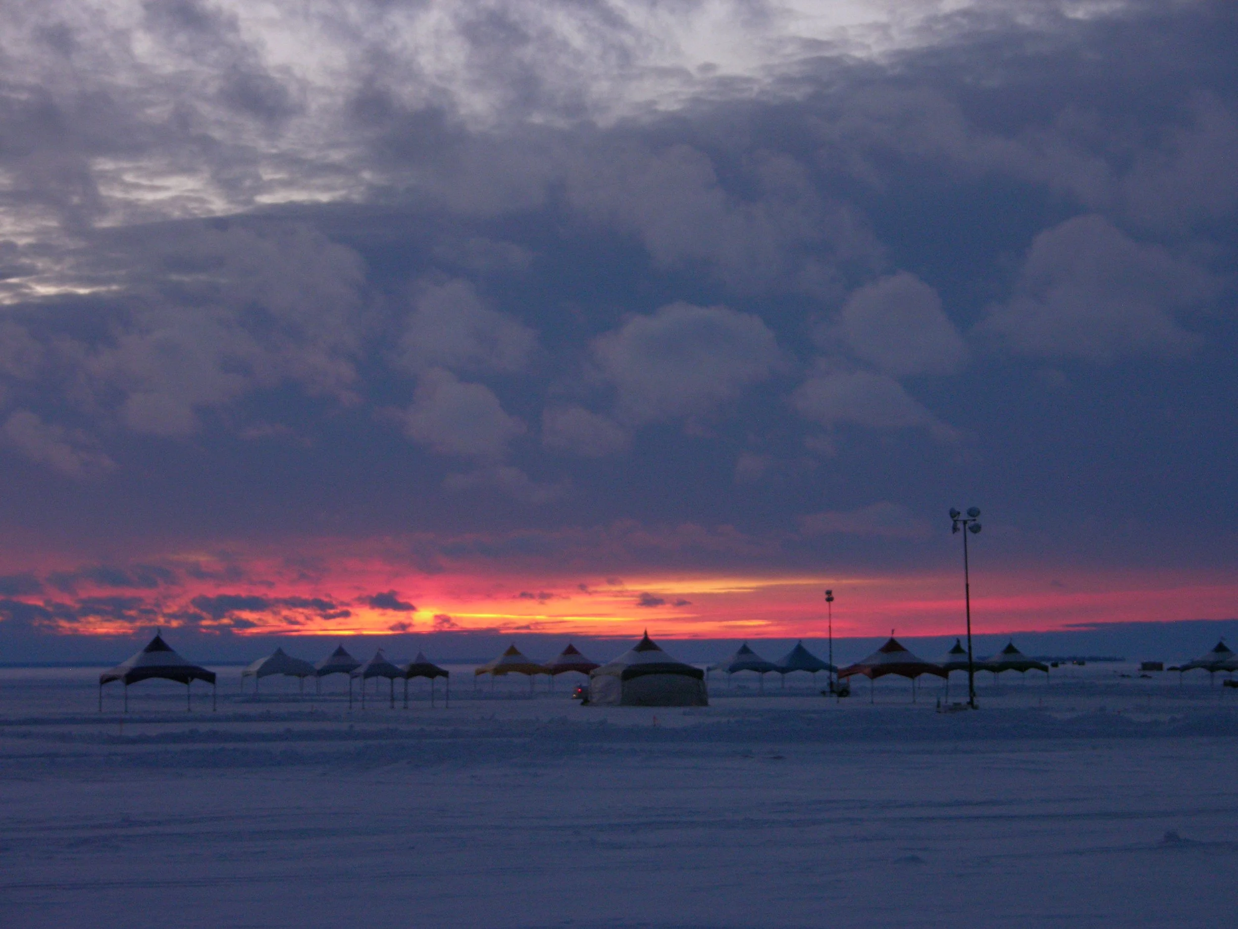 Snow-covered outdoor area with multiple tents and tall lamps under a cloudy sunset sky.