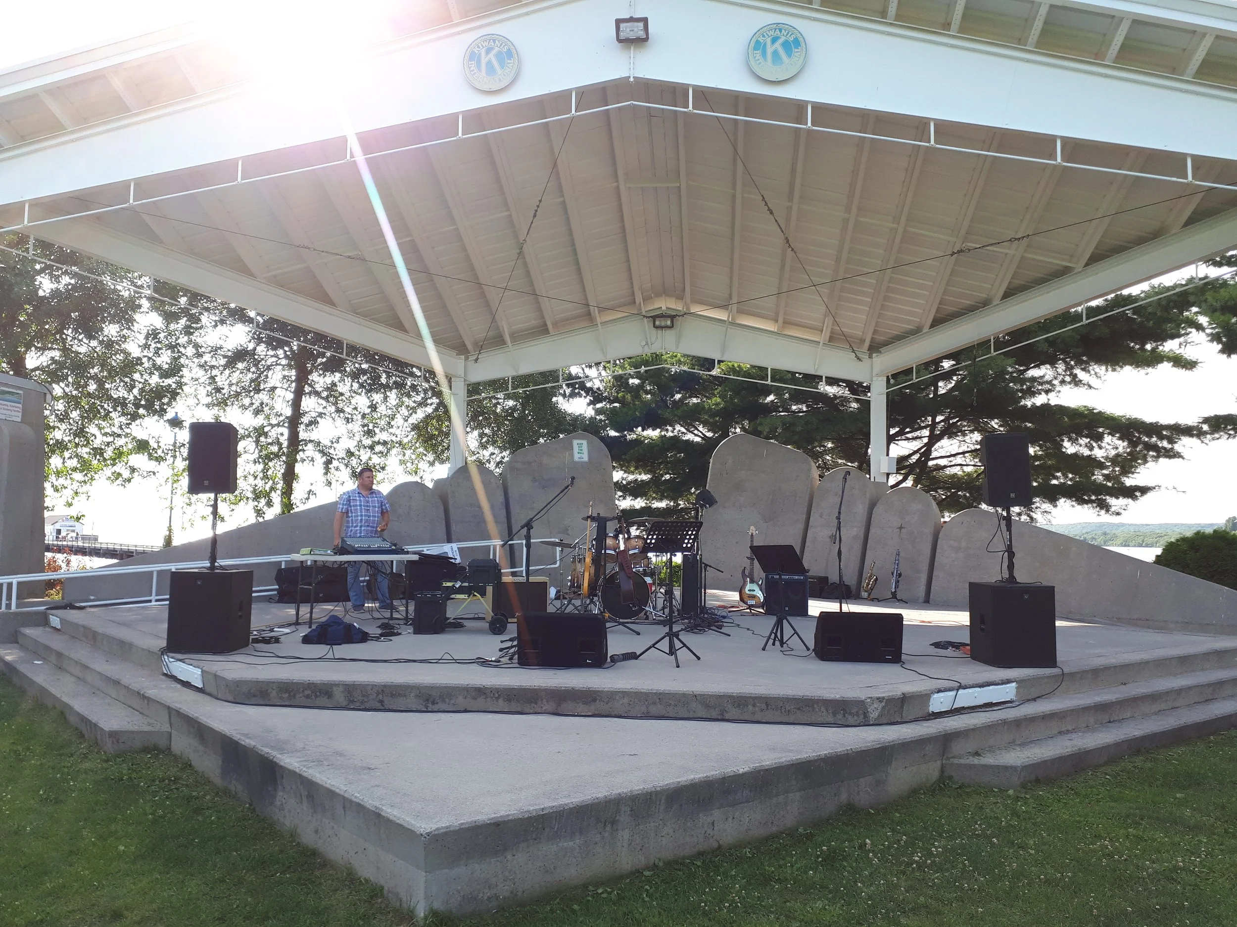 An outdoor stage setup with musical instruments and sound equipment, partially shaded by a pavilion, with a man adjusting equipment.