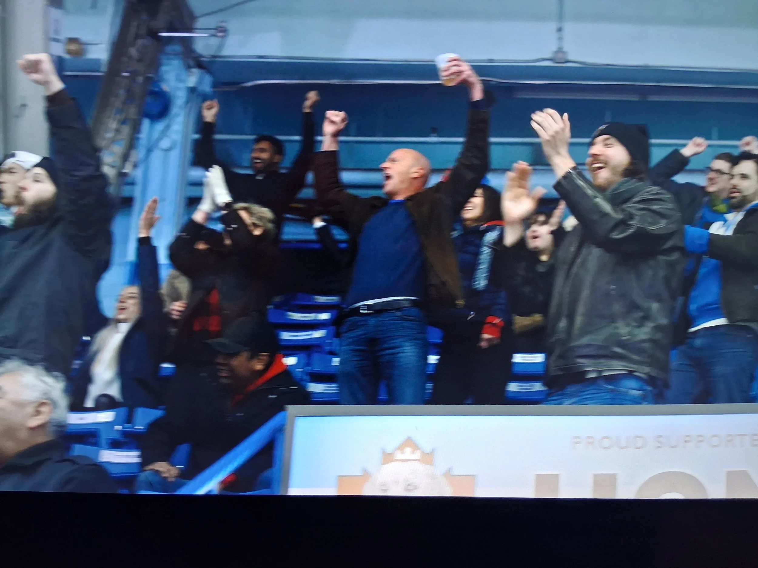 Group of people celebrating enthusiastically, some with their arms raised, in a stadium setting.