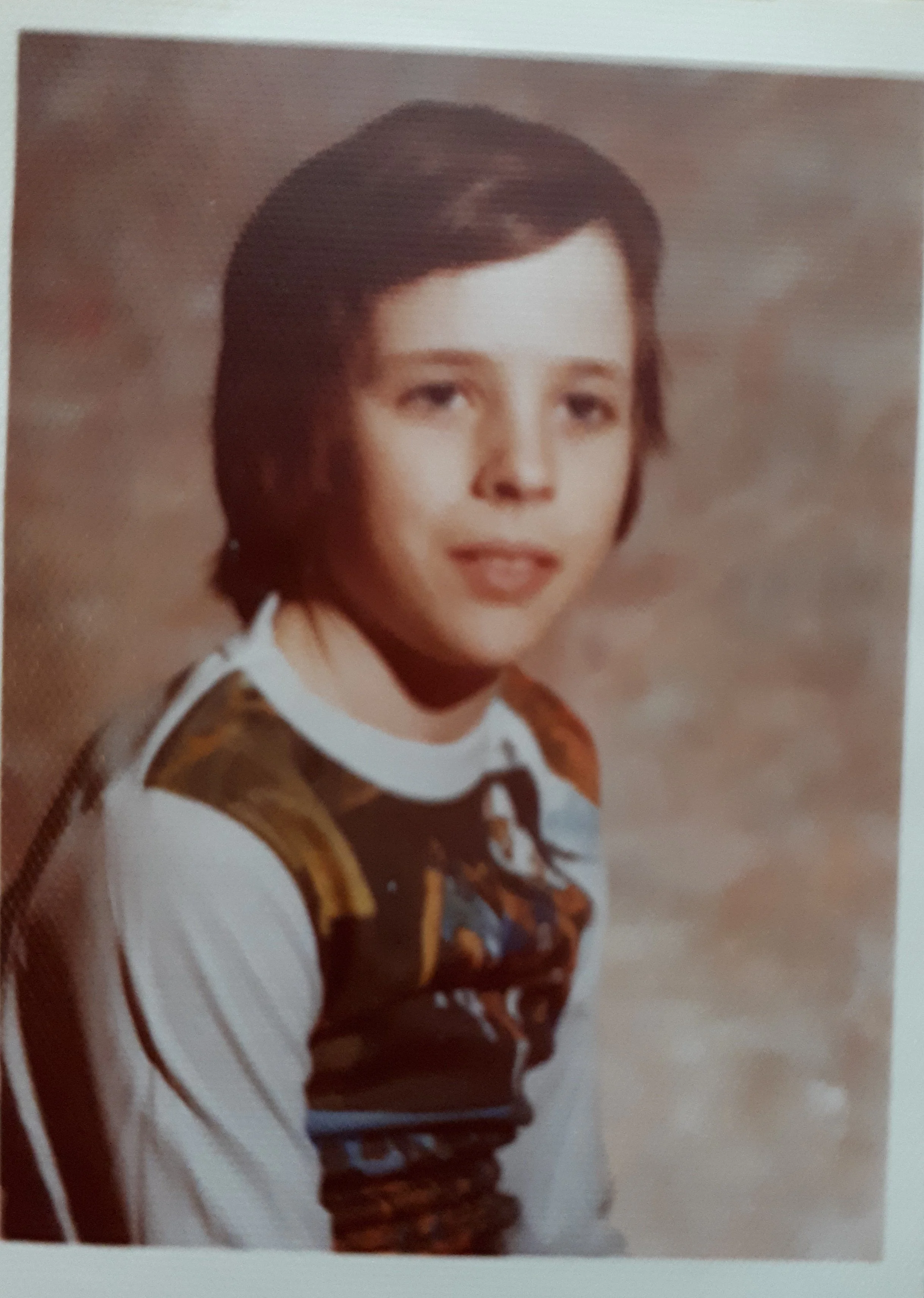 A young boy with short brown hair and a fair complexion, wearing a white shirt with a colorful vest, sitting for a school portrait against a brown and beige mottled background.