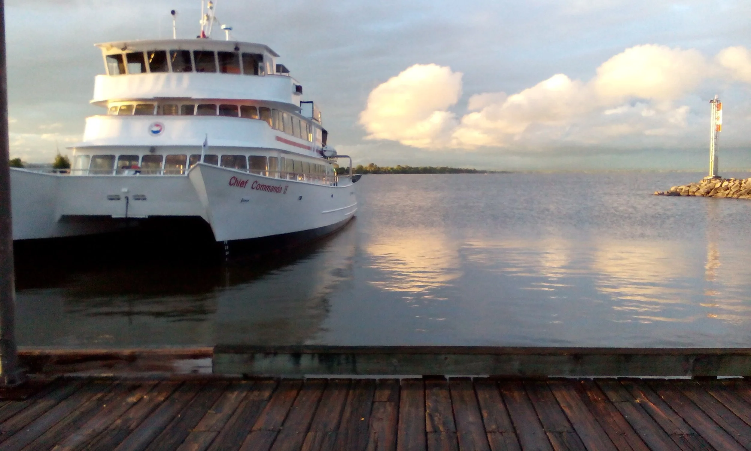 A large white boat named 'Chief Commandos II' docked at a wooden pier on a calm body of water, with a lighthouse on a small rocky jetty to the right and a partly cloudy sky in the background.