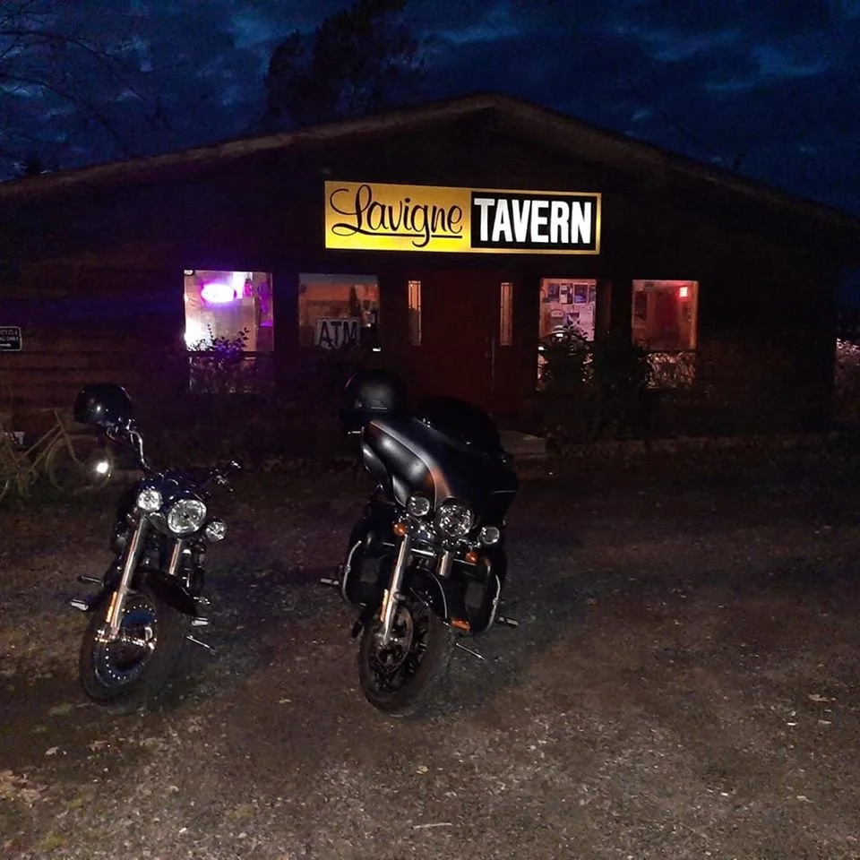 Two motorcycles parked outside the Lavigne Tavern at night, with the tavern's sign illuminated above the entrance.