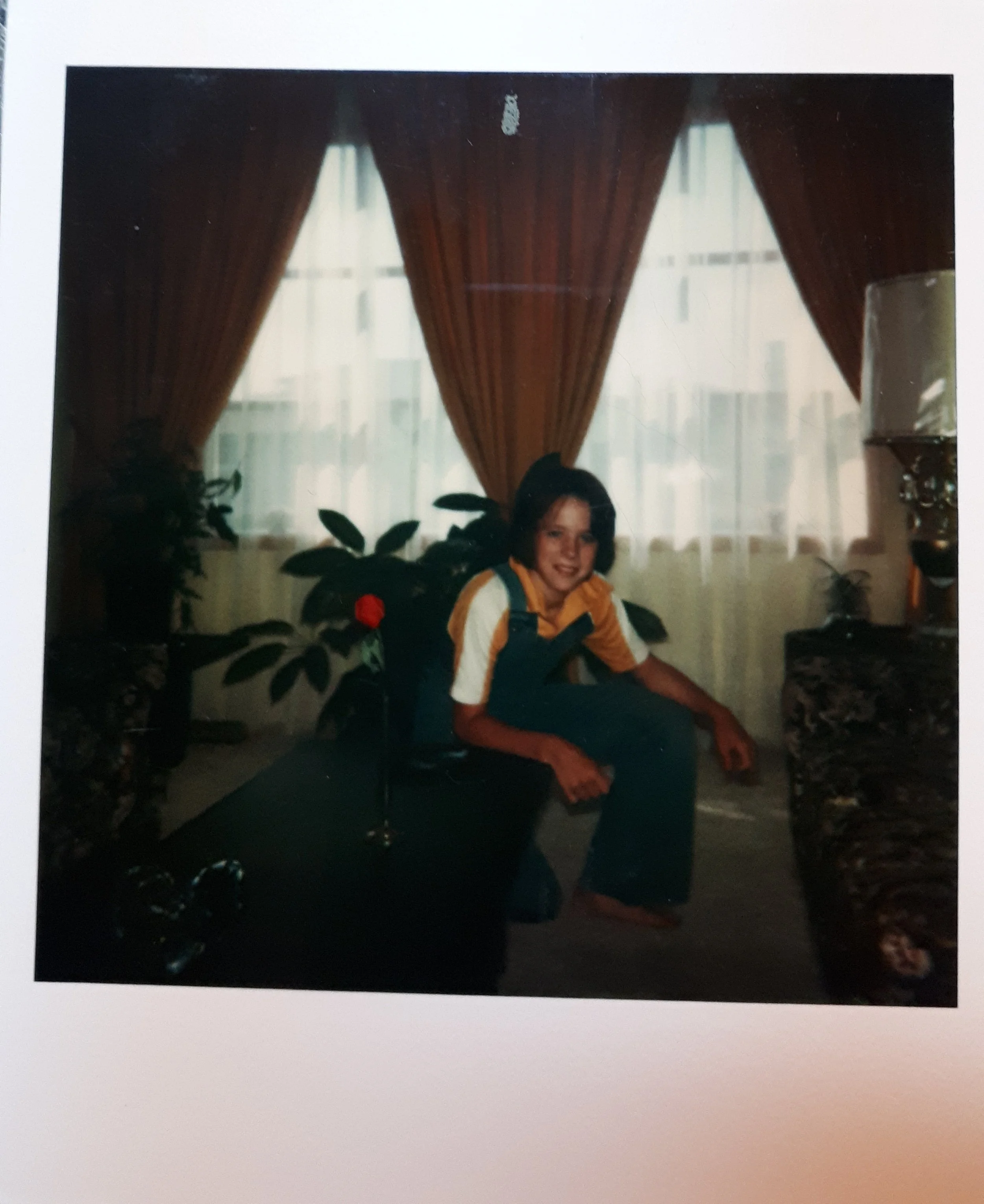 A young person with short dark hair sitting in a living room, smiling at the camera. They are wearing a yellow and white striped shirt and dark overalls. Behind them are large curtains, a window, and houseplants.
