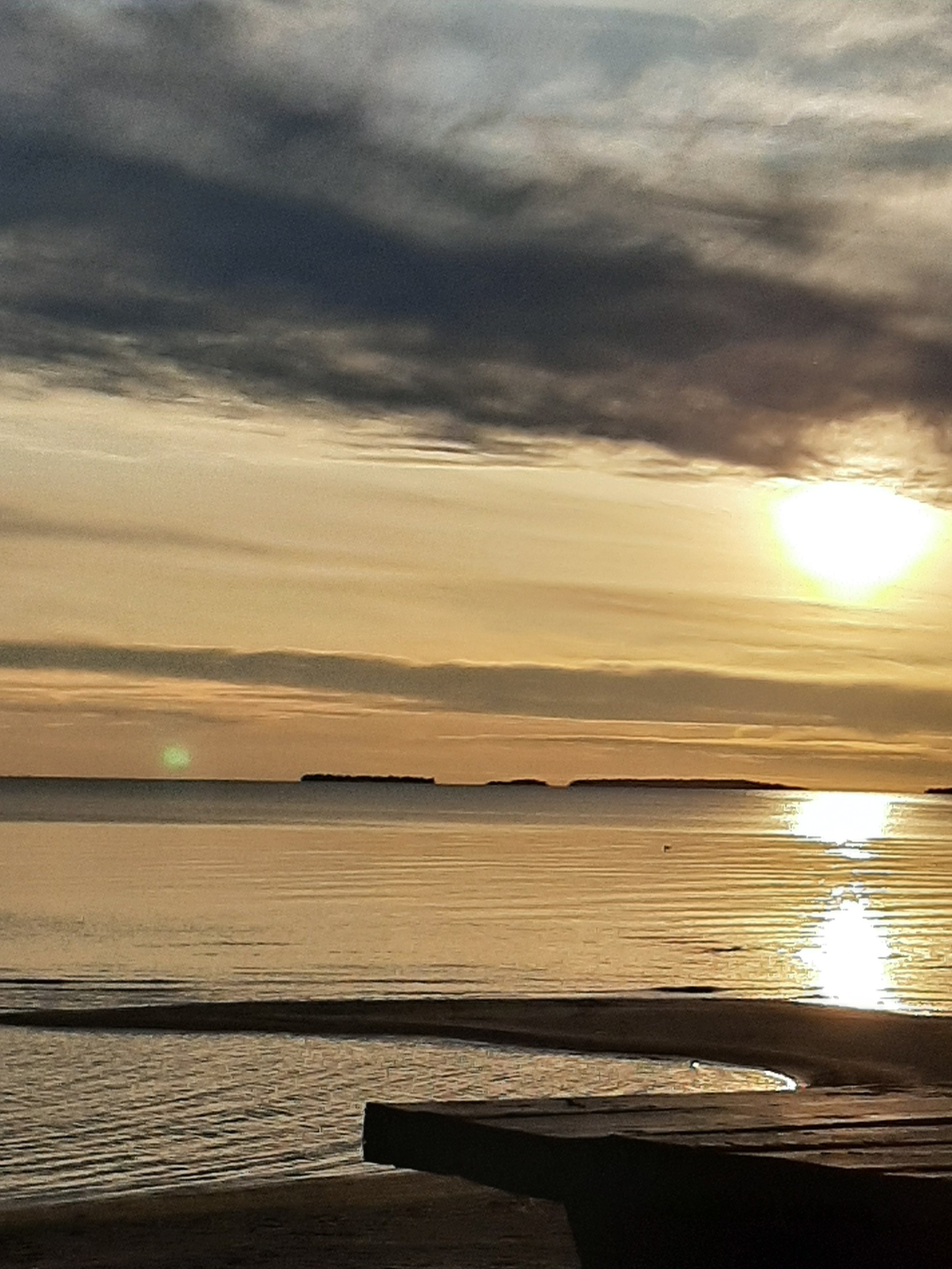 Sunset over the ocean with dark clouds in the sky and a reflection on the water, with a small landmass in the distance.