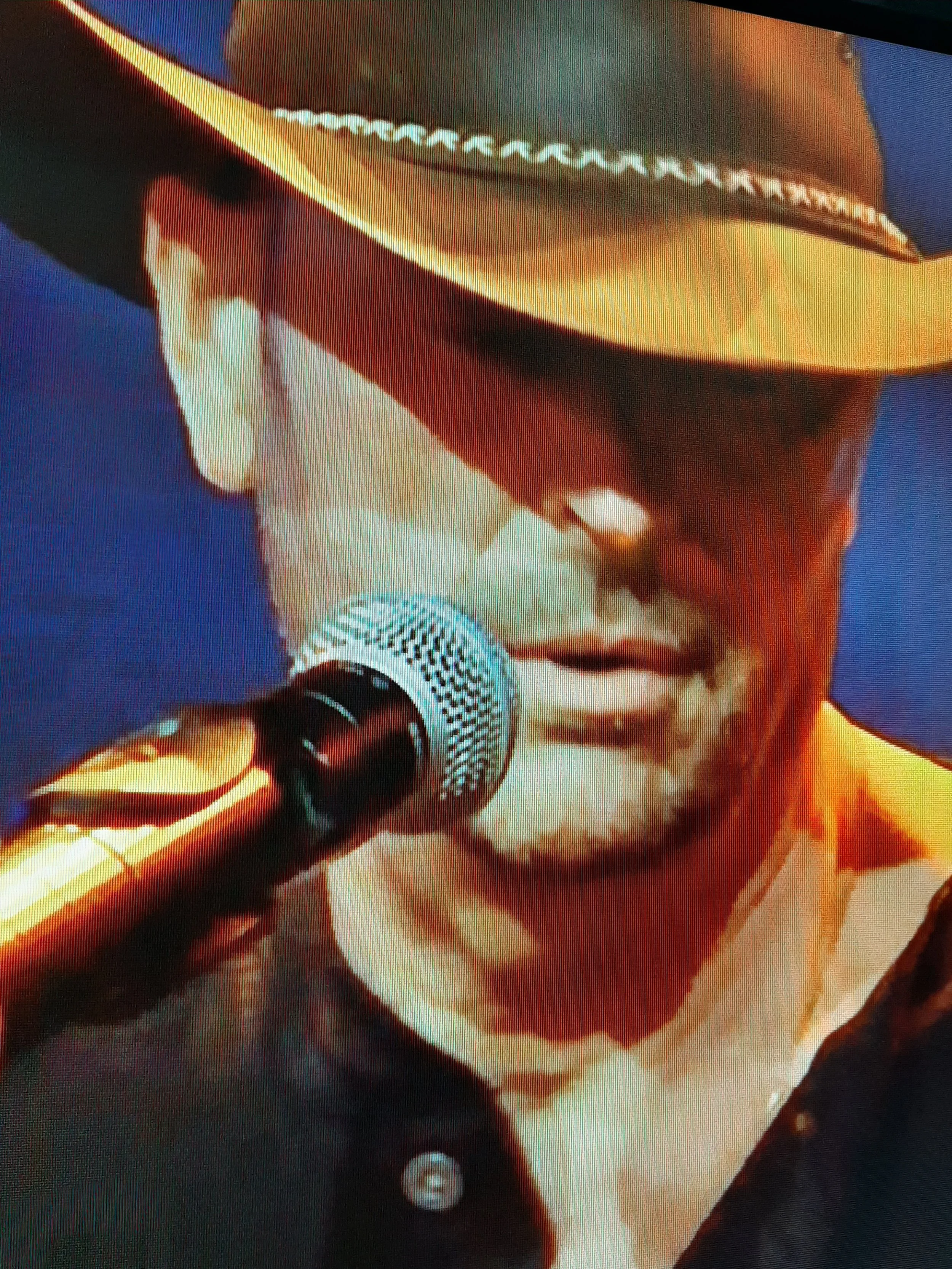 Close-up of a man wearing a cowboy hat and sunglasses speaking into a microphone.