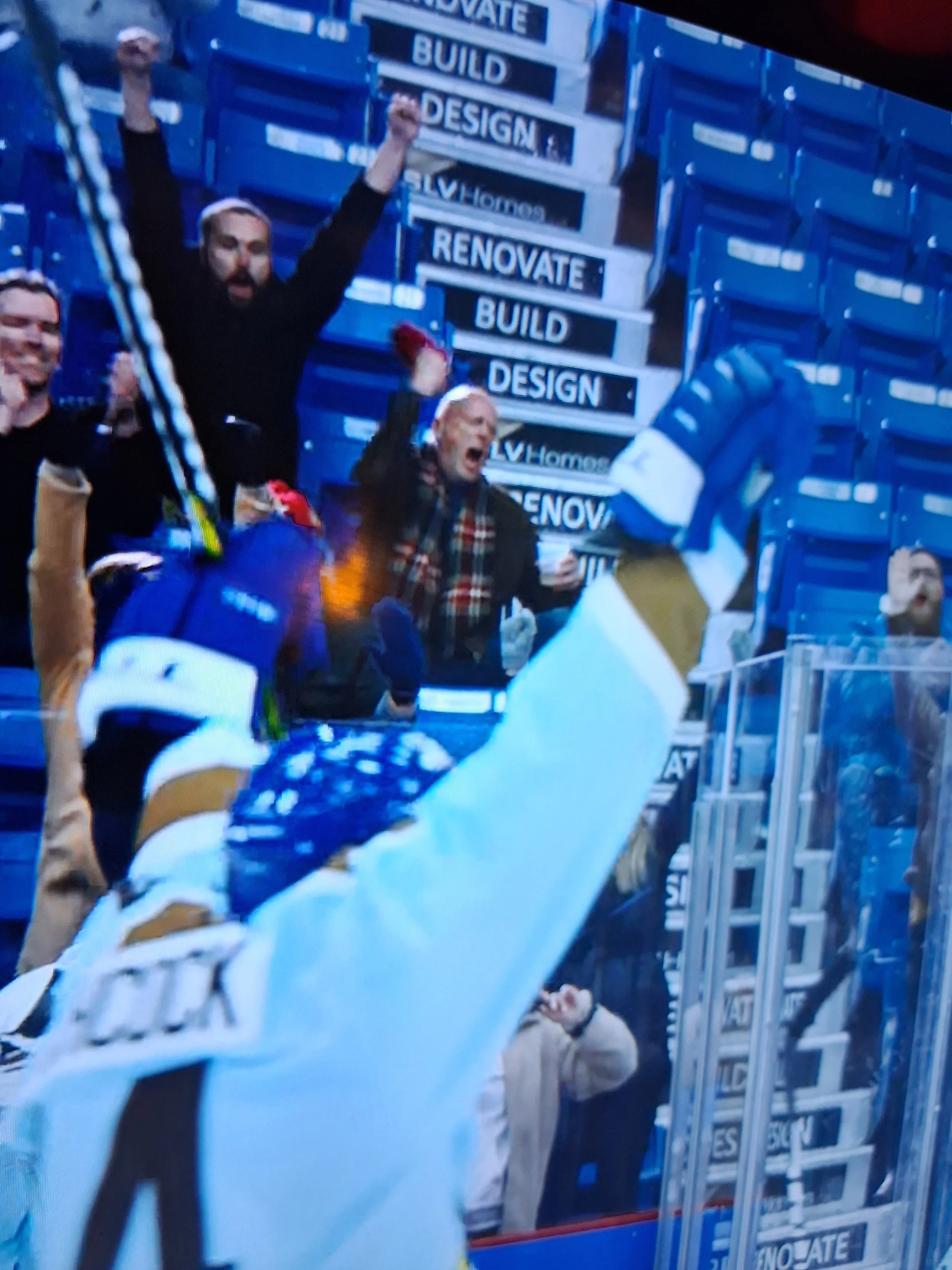 Celebrating hockey players and fans on the ice with raised fists and cheering, with a background of blue signs that say 'BUILD,' 'RENOVATE,' 'DESIGN,' and 'HOME.'