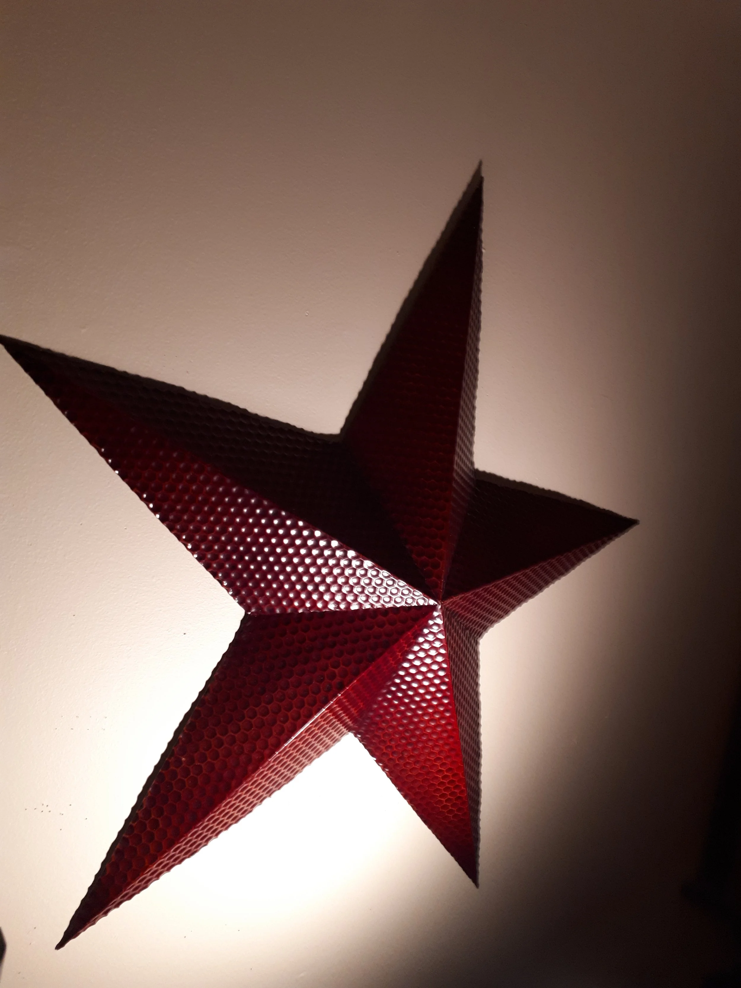A three-dimensional red star decoration with a textured surface, hanging against a plain wall with lighting creating shadows.