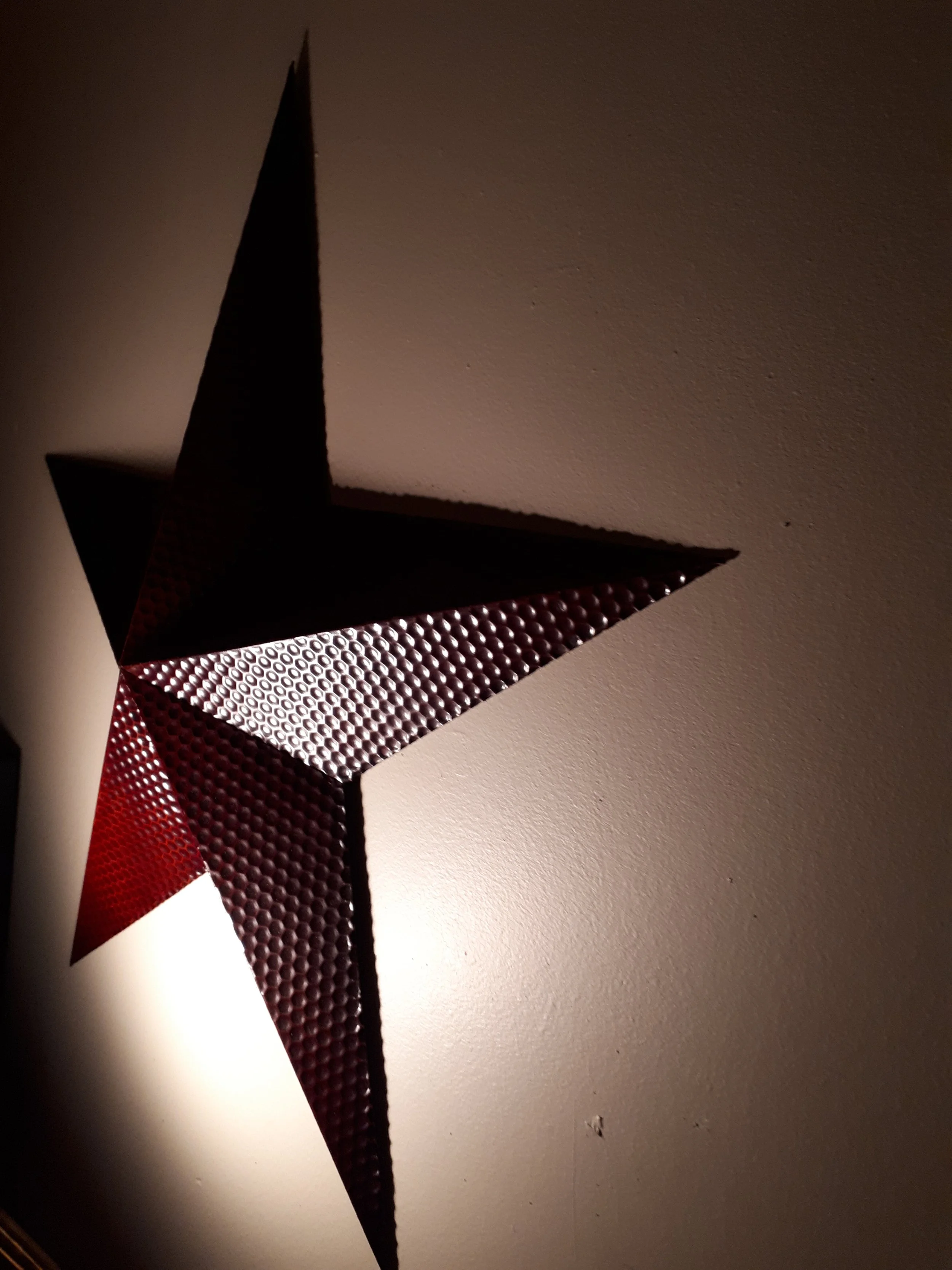 Decorative star-shaped wall hanging with textured metallic surface, illuminated from below, casting a shadow on the wall.