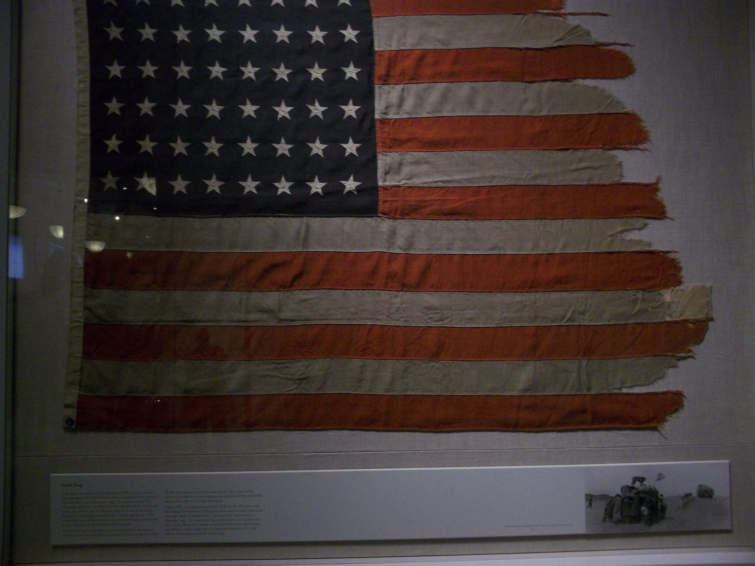 An American flag with worn, tattered fabric, displayed in a museum setting.