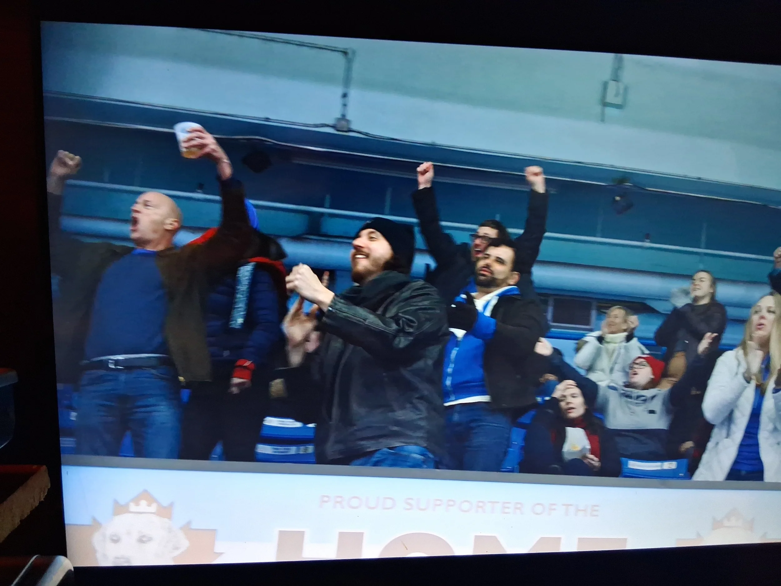 A group of sports fans, cheering and celebrating, with some raising their fists or drinks, in a stadium setting.