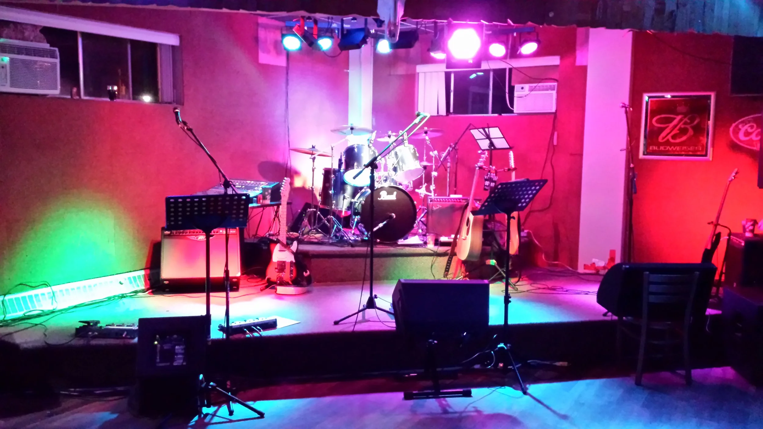 Stage with musical instruments including drums, guitars, and microphones under colorful stage lights in a music venue or bar.