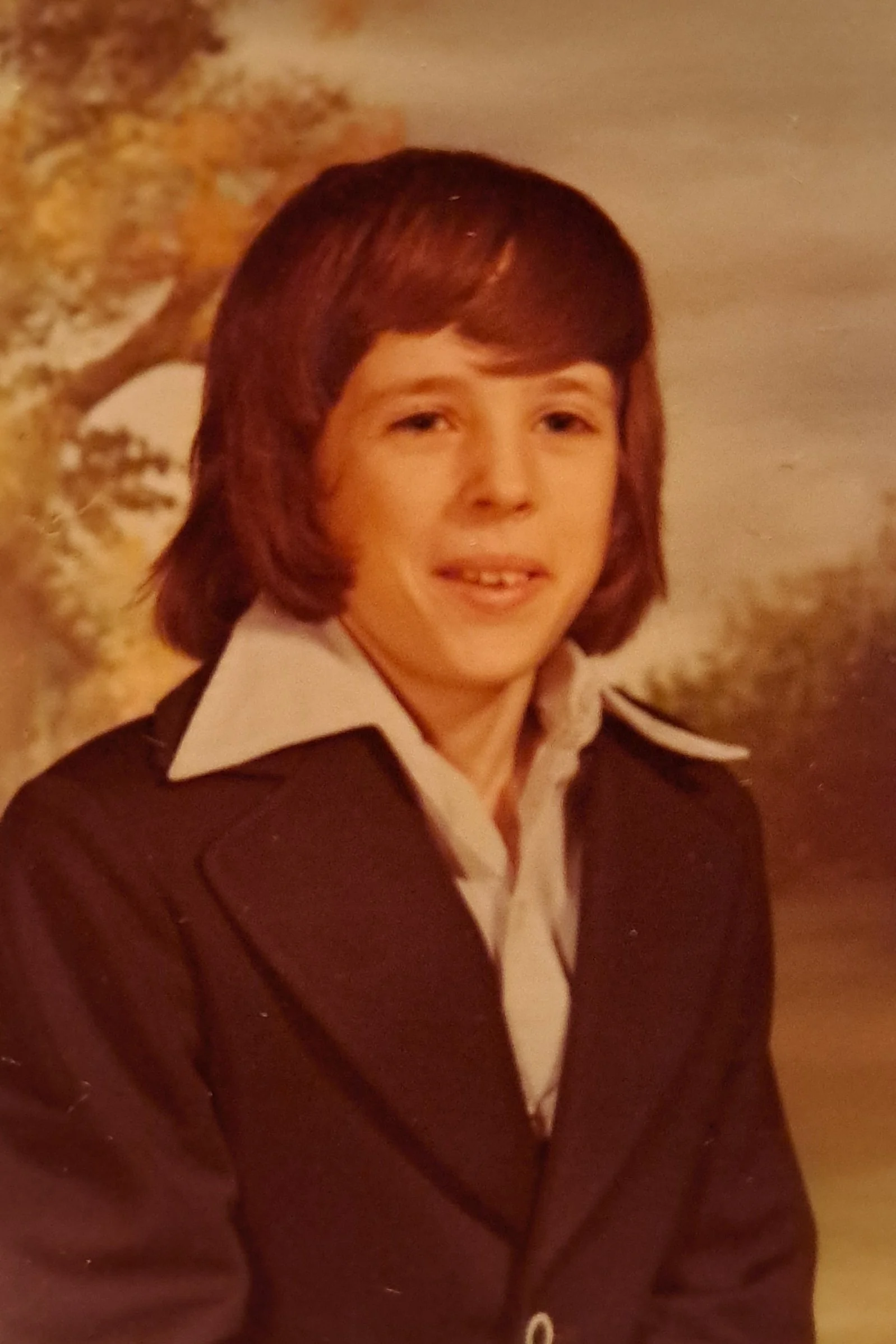 A young boy with medium-length brown hair wearing a white collared shirt and a brown blazer, outdoors with trees in the background.