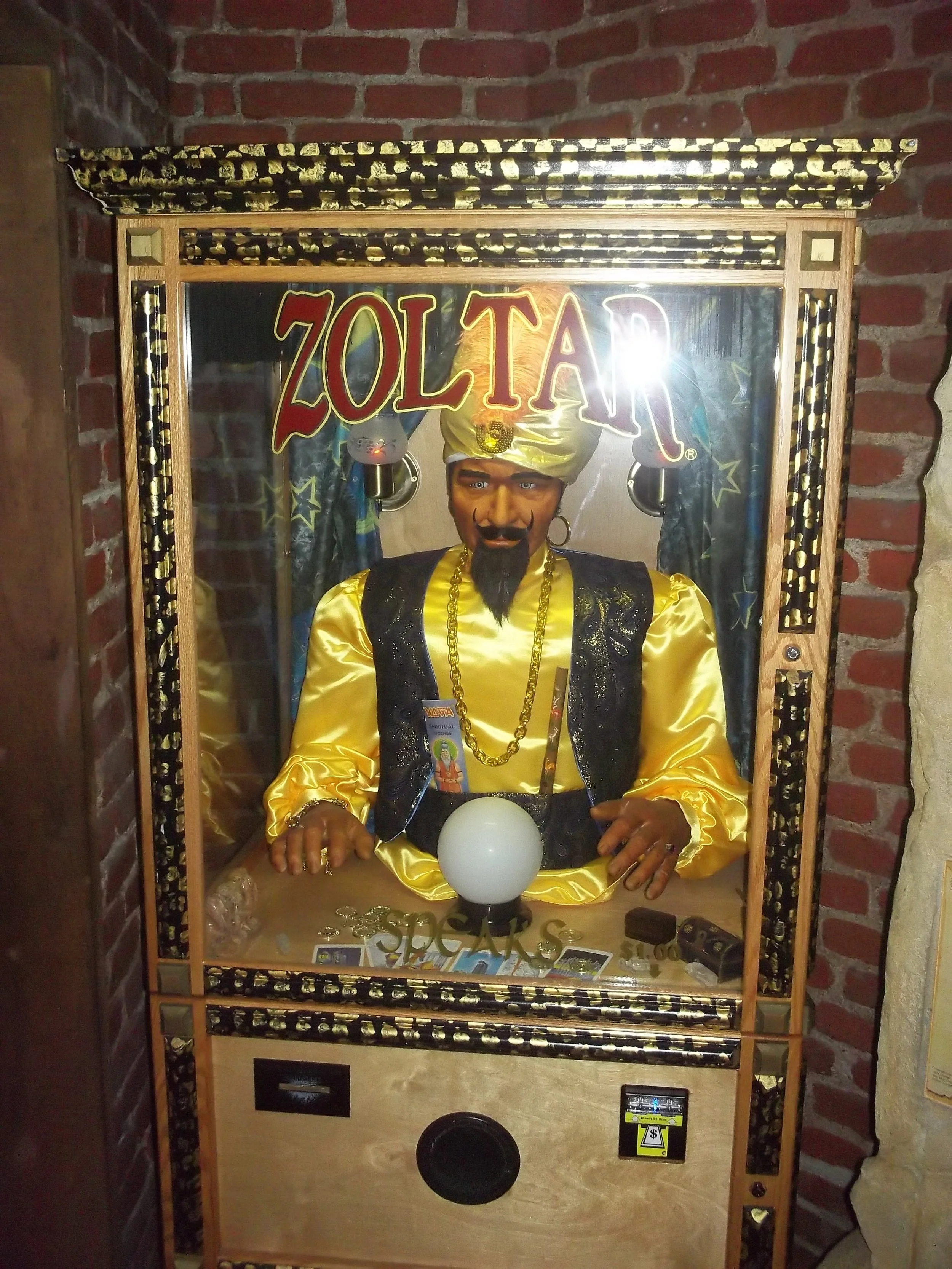 A Hanuman statue dressed in yellow and black with gold jewelry, inside a glass display case with a red brick wall background, and the name 'ZOLTAR' in red and yellow letters at the top.