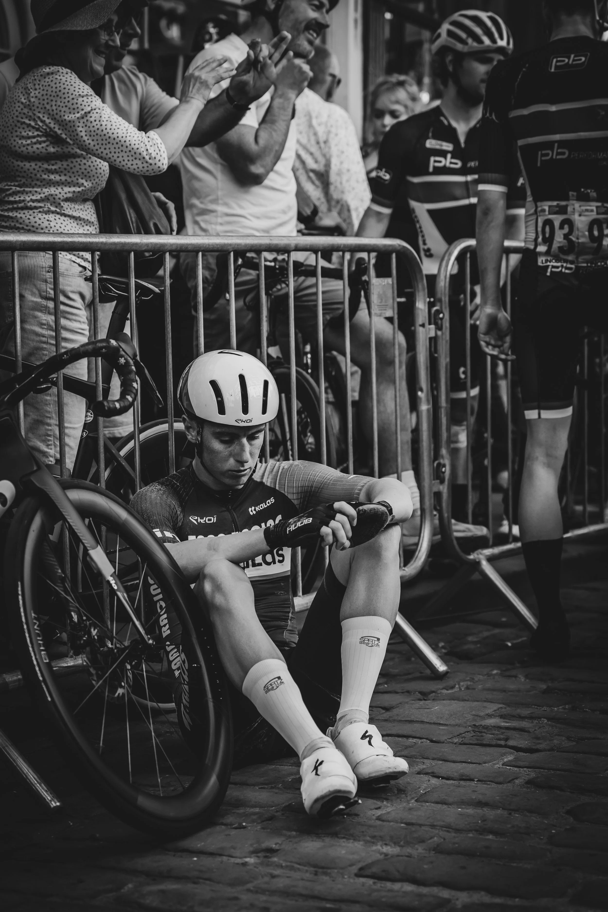 A cyclist wearing a helmet sits on the ground with their head bowed and their bicycle fallen beside them, while spectators and other cyclists stand behind a barrier, some making gestures and smiles.