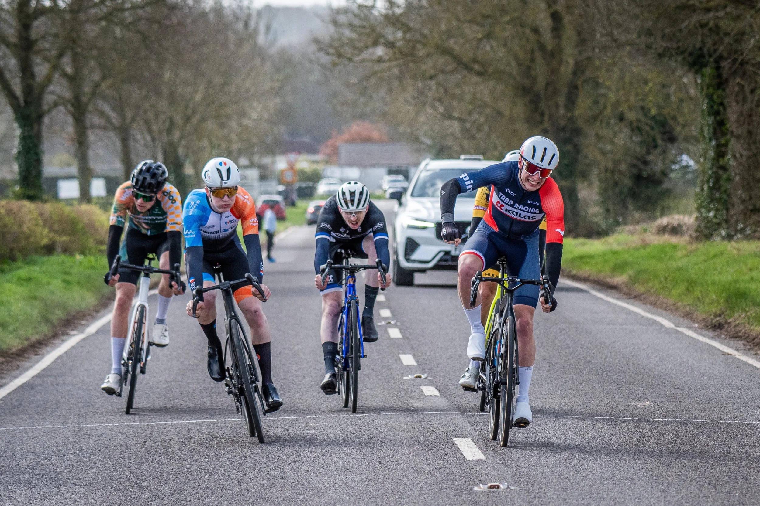 Cyclists racing on a rural road, with one cyclist crossing a line on the asphalt, while other riders pedal behind him. Car is visible in the background, trees lining the road, and overcast sky.
