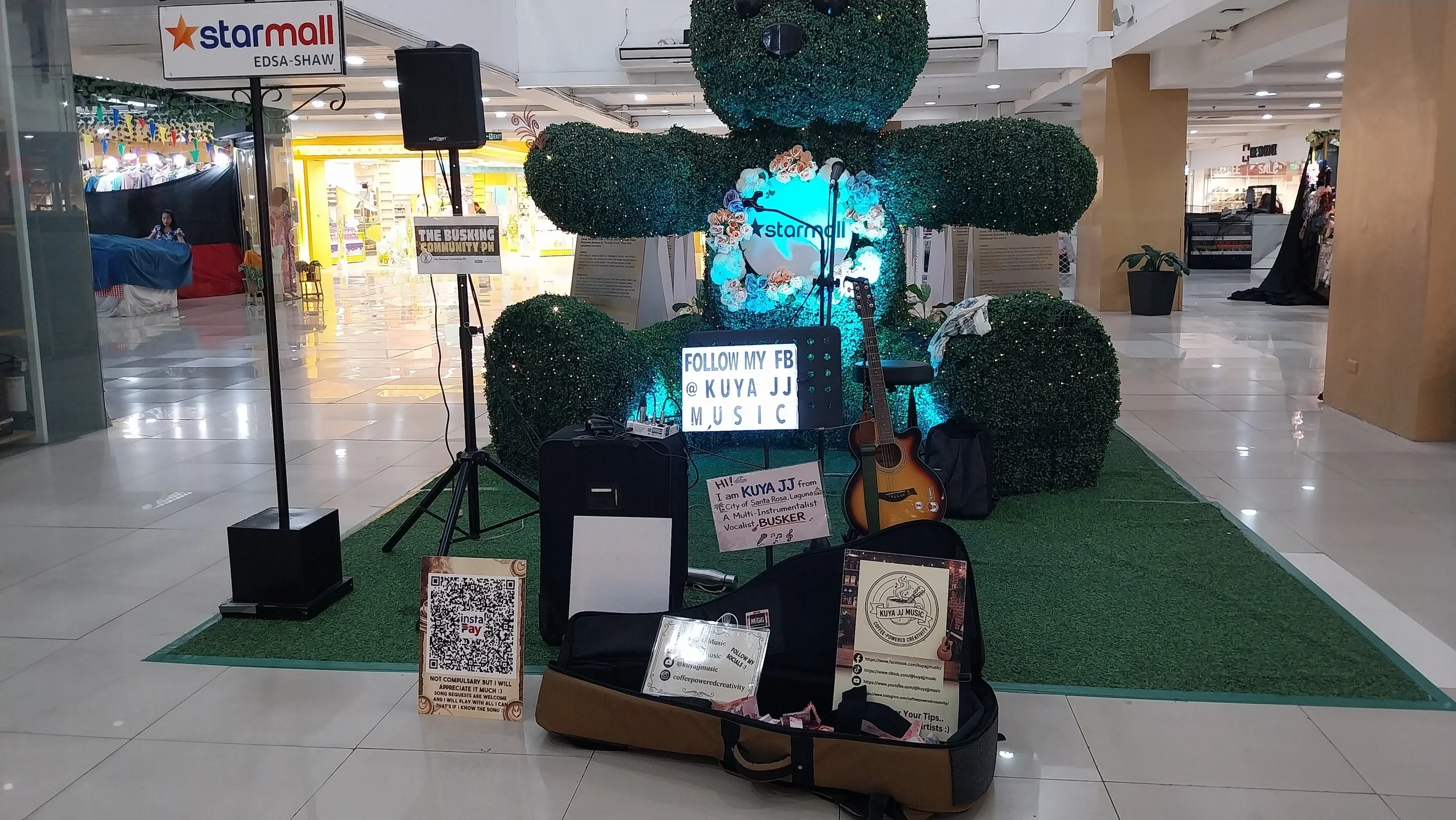 A small music setup in a shopping mall, featuring a green hedge backdrop with the star mall logo, an acoustic guitar, a microphone, and signage encouraging following a Facebook page. There are various promotional signs, a QR code, and some audio equi