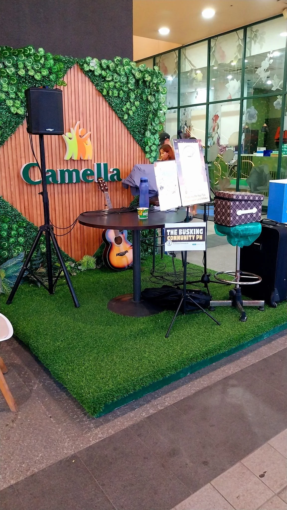 A small outdoor stage setup with a guitar, speaker, and music stand, beneath a decorative green foliage backdrop with the word "Camella" and a logo, in front of a glass building with floral patterns.