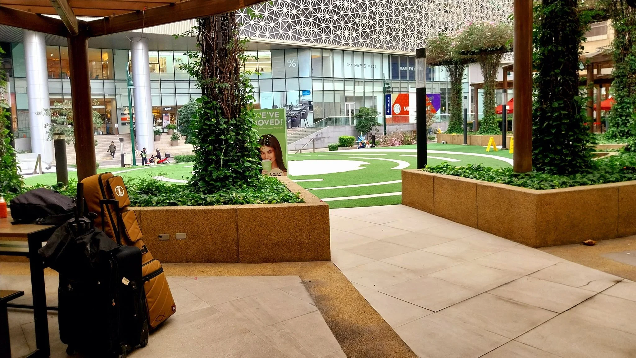 Entergarden area with potted plants and trees in a city setting, with a grassy courtyard and modern buildings in the background, luggage at a table, and a poster showing a woman drinking coffee.