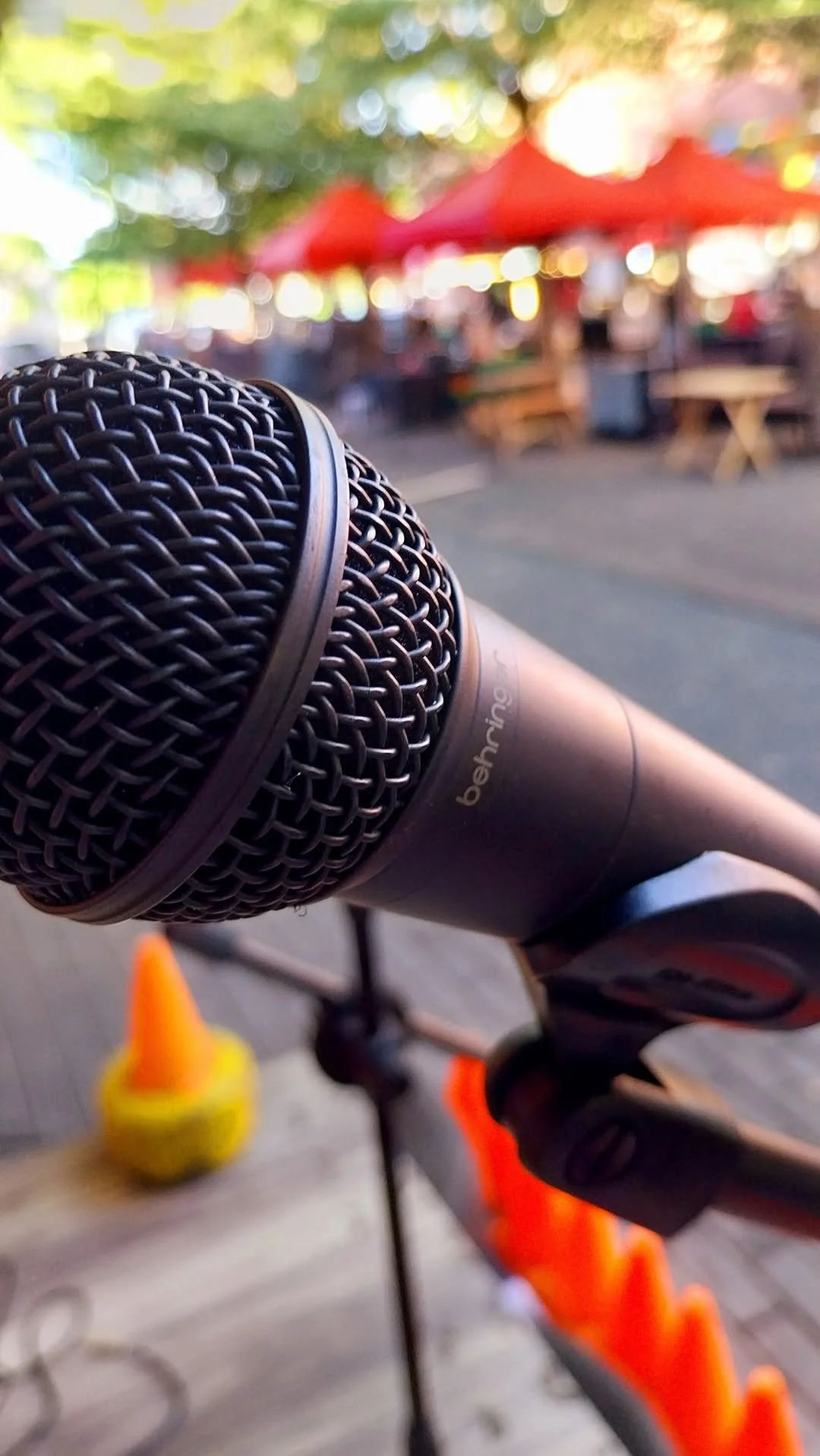 Close-up of a Benro microphone mounted on a bicycle handlebar, with blurred background showing outdoor seating with red umbrellas and trees.