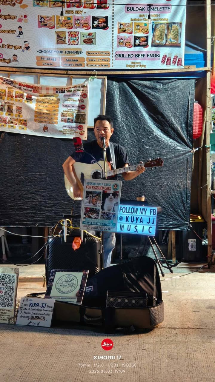 A male musician with a guitar singing into a microphone at an outdoor event. Behind him are menu signs for food items like buldak omelette and grilled beef enoki, with some promotional and social media signs. There is a black backdrop, a small amplif