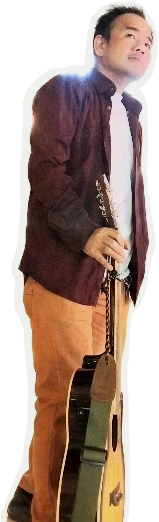 A man holding an acoustic guitar, standing with one hand in his pocket, wearing a dark jacket and beige pants, with a light background.